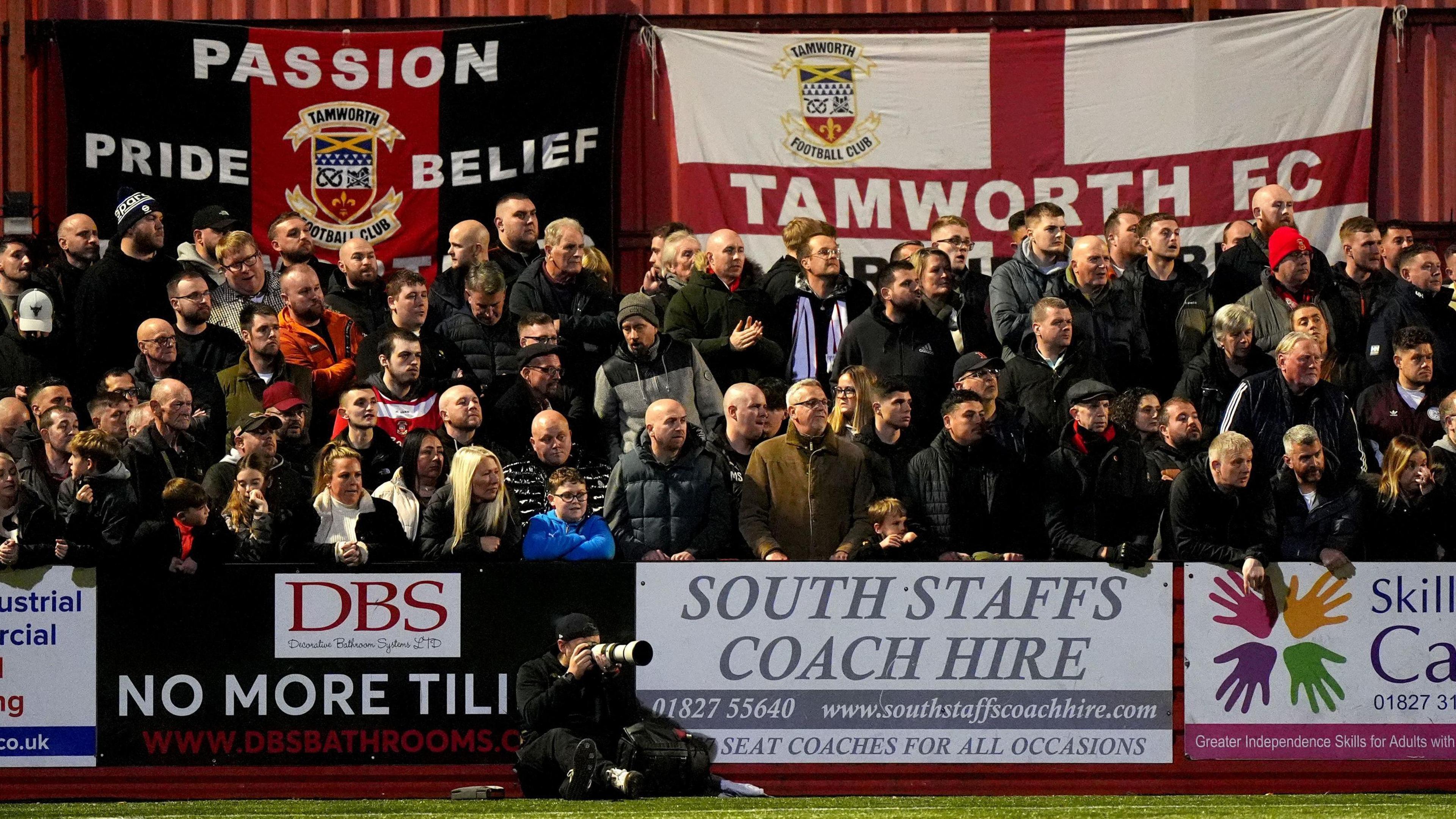 Tamworth fans in the stands. There are sponsorship boards around them and flags in the background. 