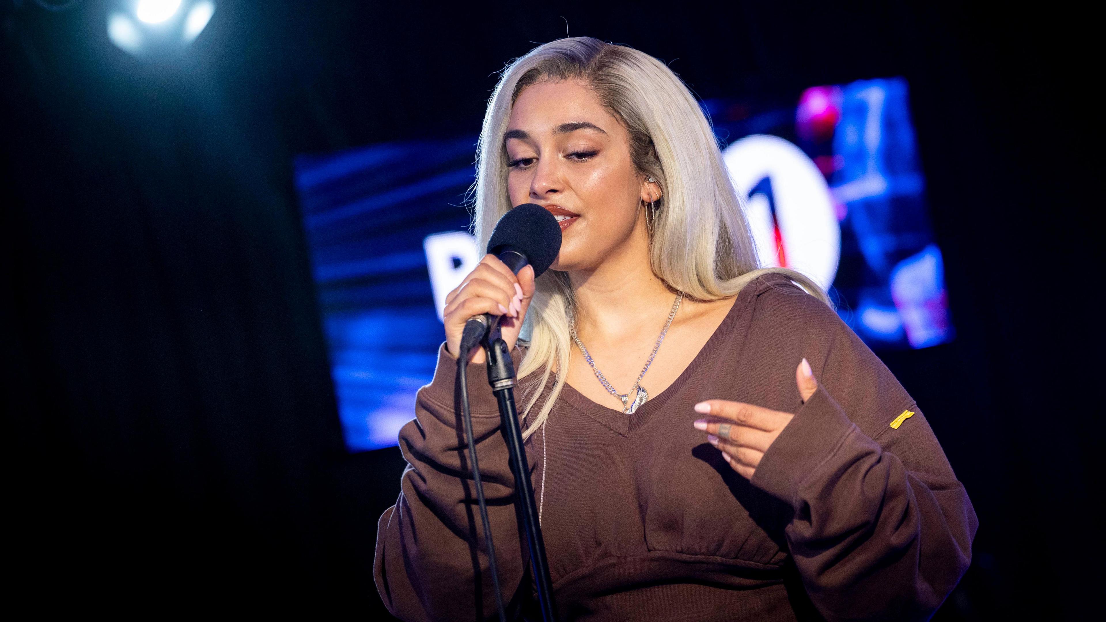 Jorja Smith playing at Radio 1's Live Lounge in March 2025. She is in a brown, loose fitting top and has blonde hair. She is singing into a microphone in a dark room.