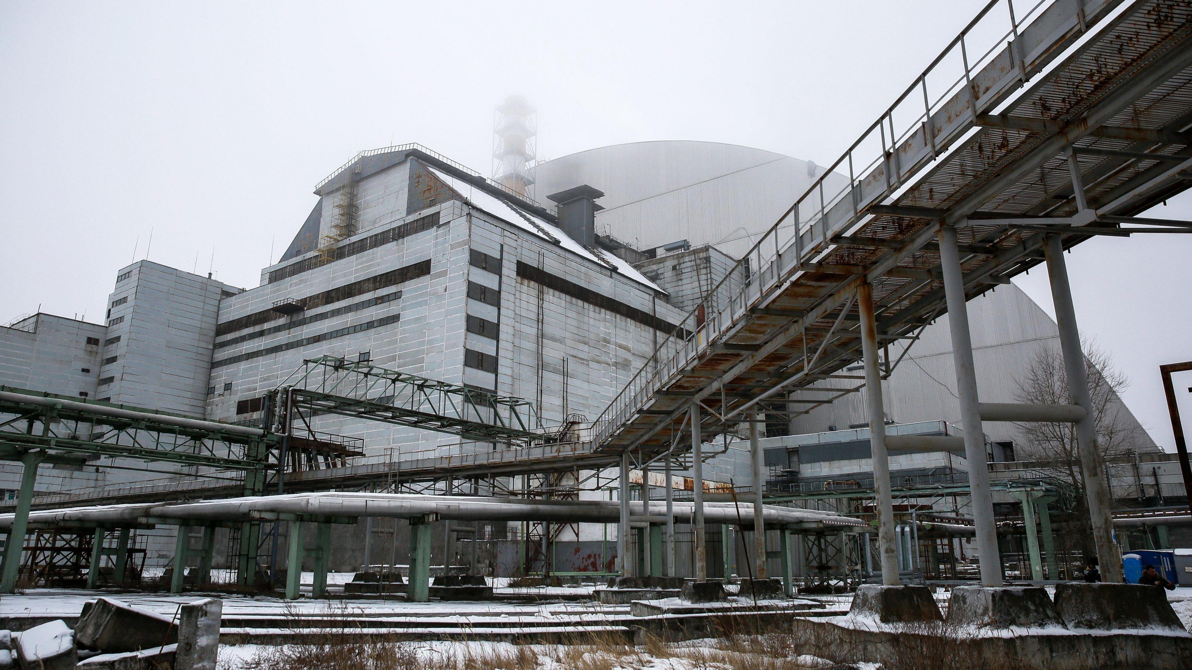 A grey industrial building with various pipes, walkways and platforms attached to it stands in front of a large, dome-shaped structure of a similar colour. The ground is covered with snow and the metallic structures show signs of rust and age.