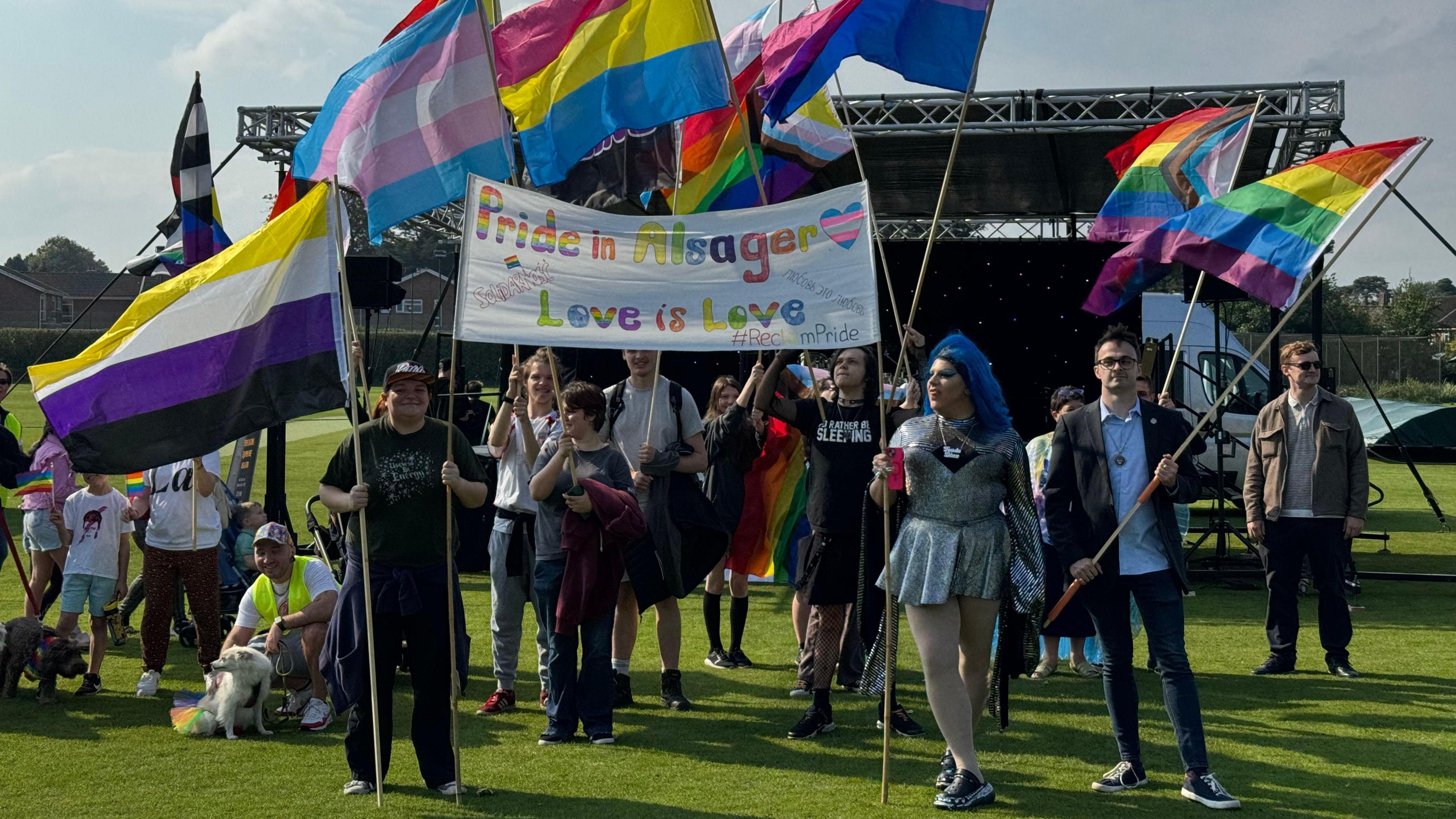 A wide variety of colourful flags representing different aspects of the LGBT community are being held by a group of people stood on a grass cricket pitch with a stage in the background.