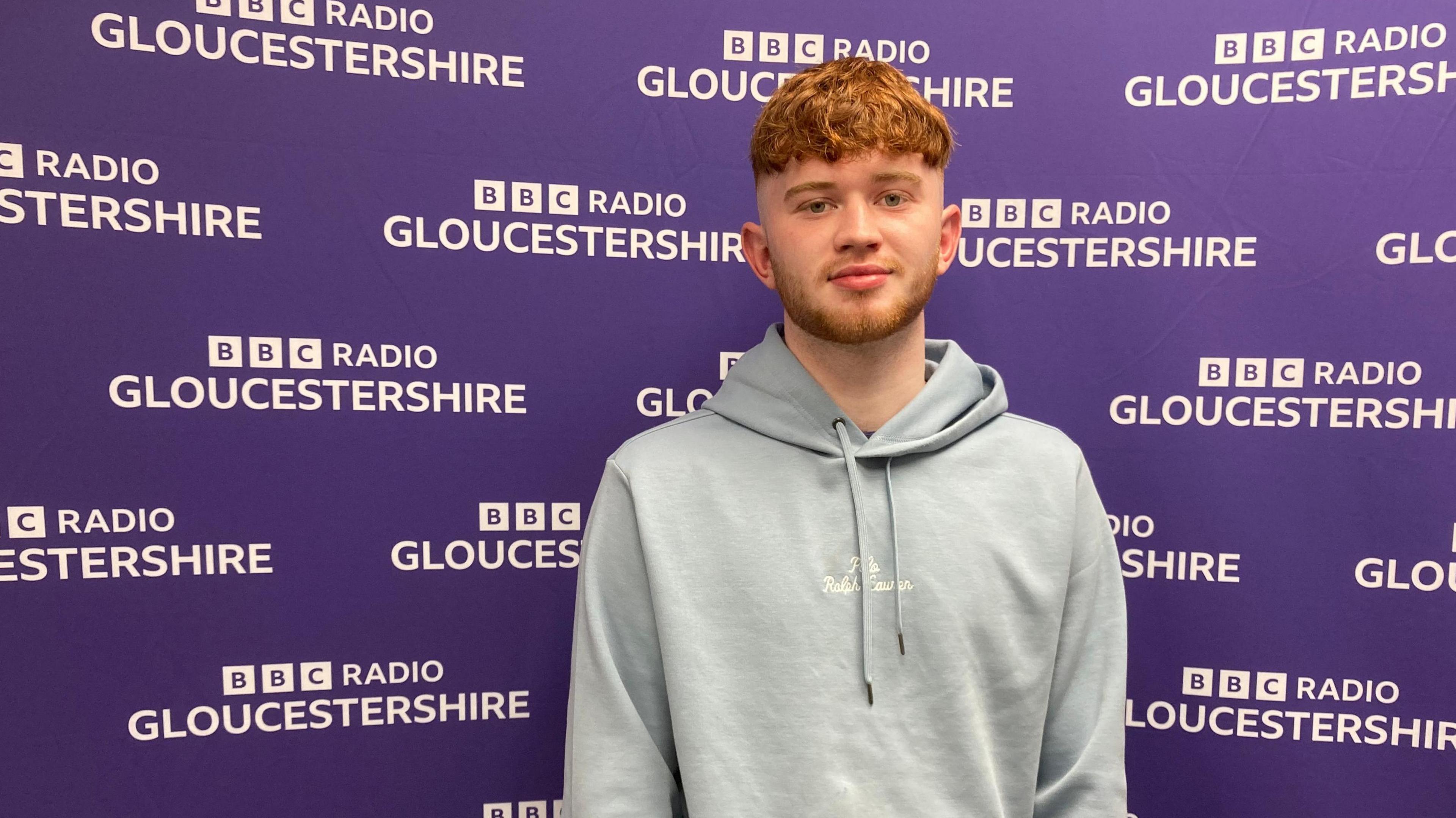 A ginger-haired young man in a grey hoodie standing in front of a purple BBC Radio Gloucestershire branded backdrop with white writing on it