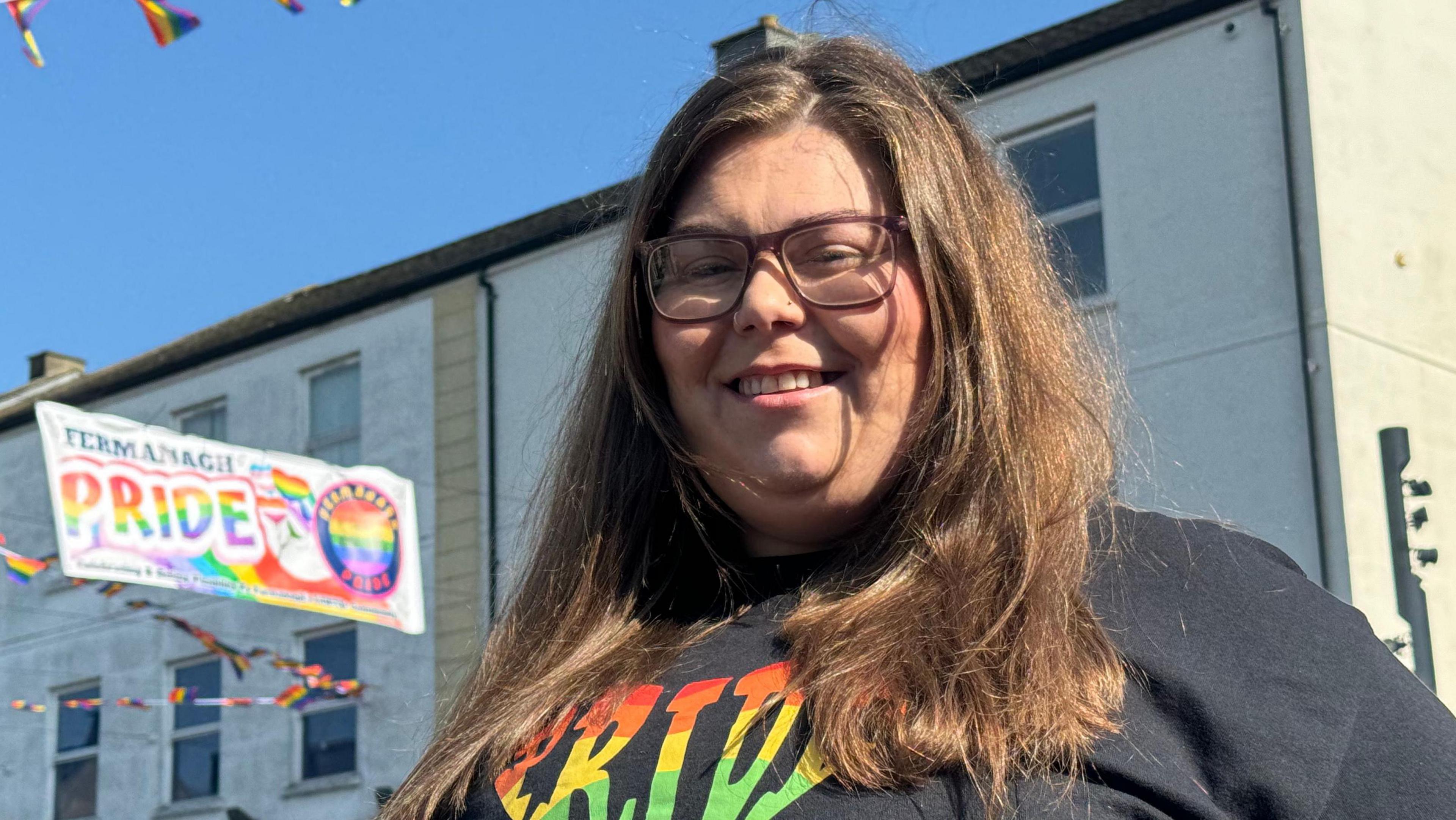 Geraldine has long brown hair and wears glasses and has a pride top on. Hanging up in the street is a sign for Fermanagh pride. 