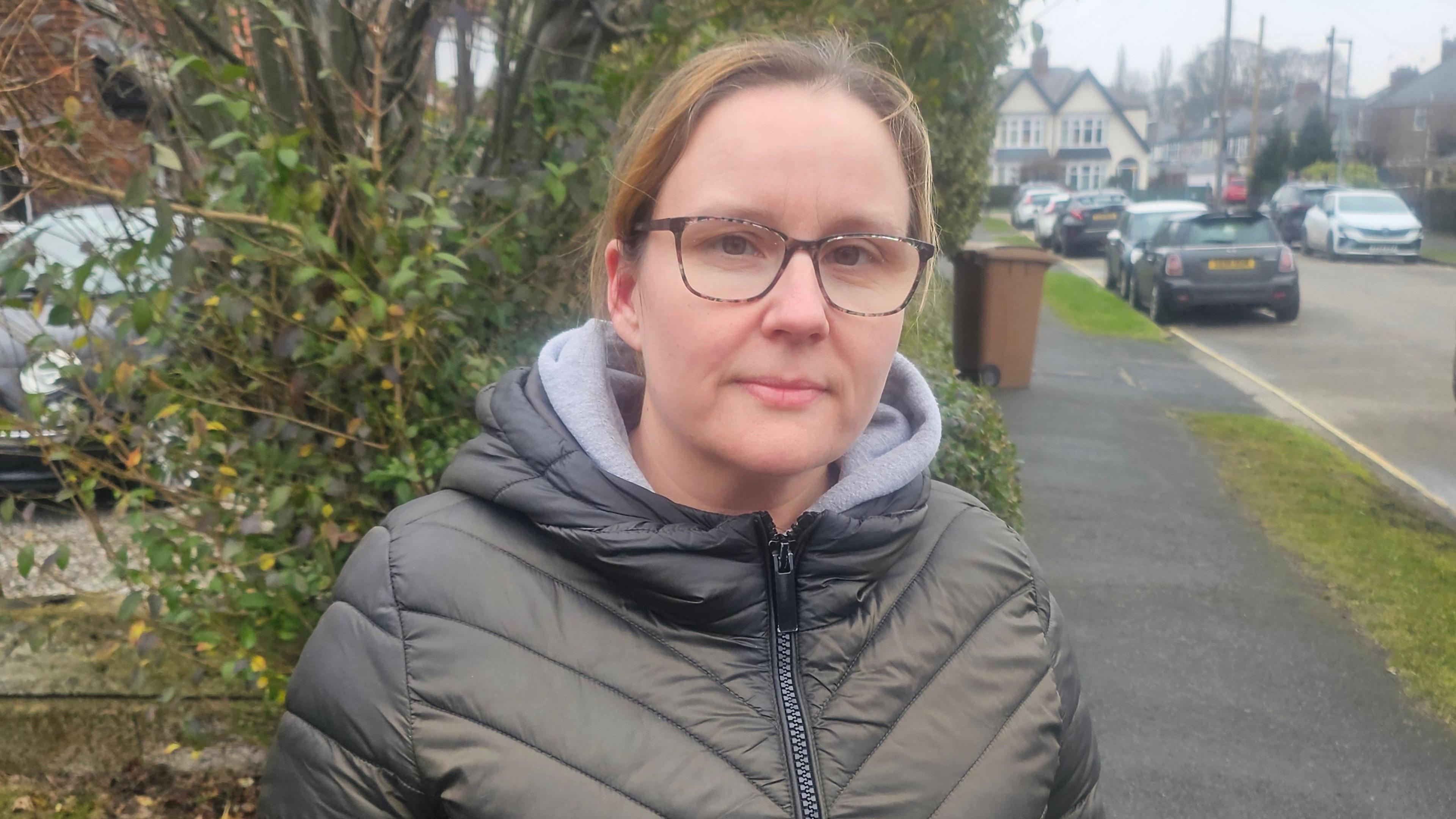 A woman stands at the side of a road which has cars parked along a grass verge. She is wearing dark rimmed glasses and a green quilted jacket.