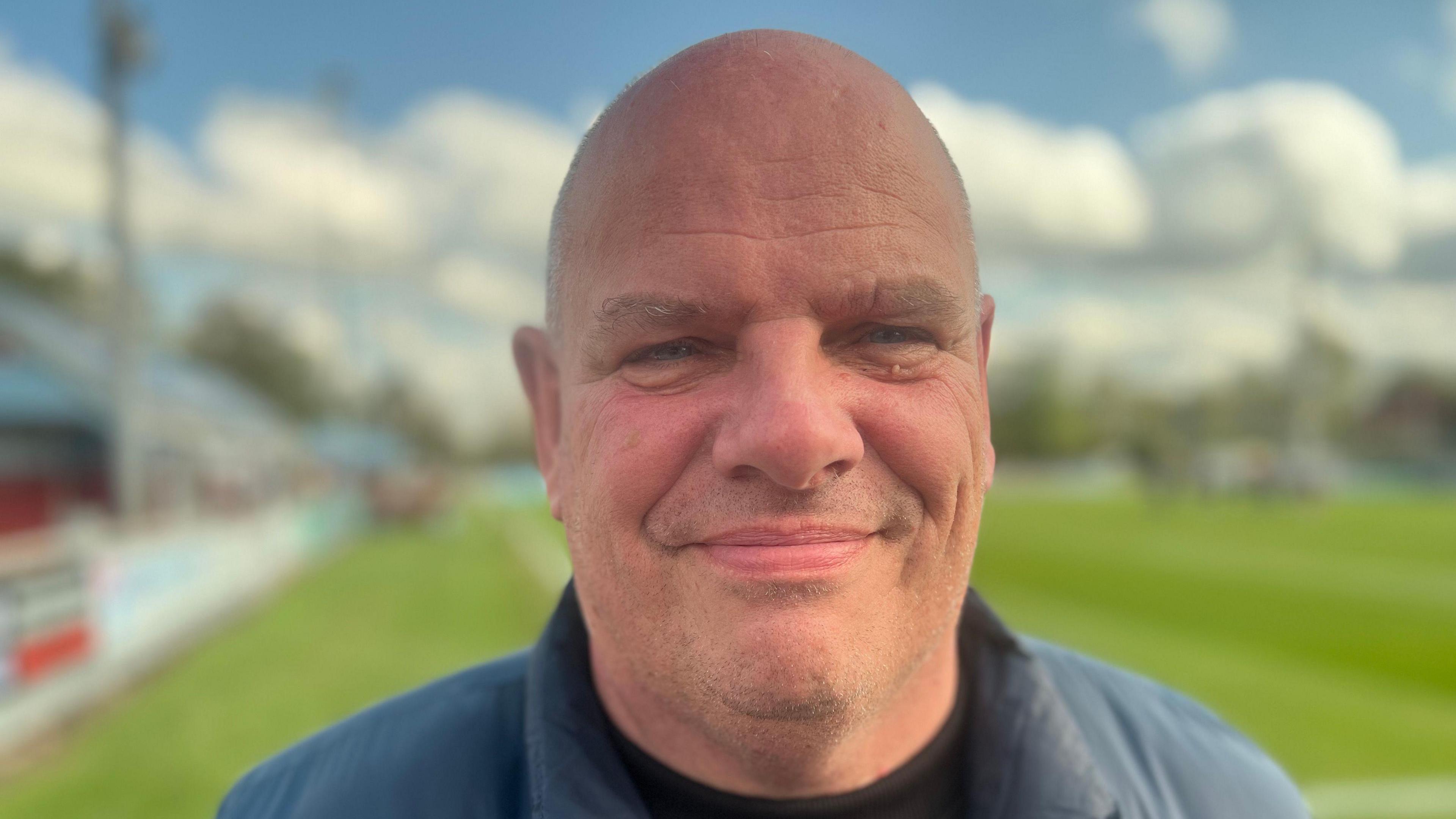 Karl Lindsay looks into the camera with a small smile, he is bald and wears a blue jacket, with the football pitch blurred in the background.