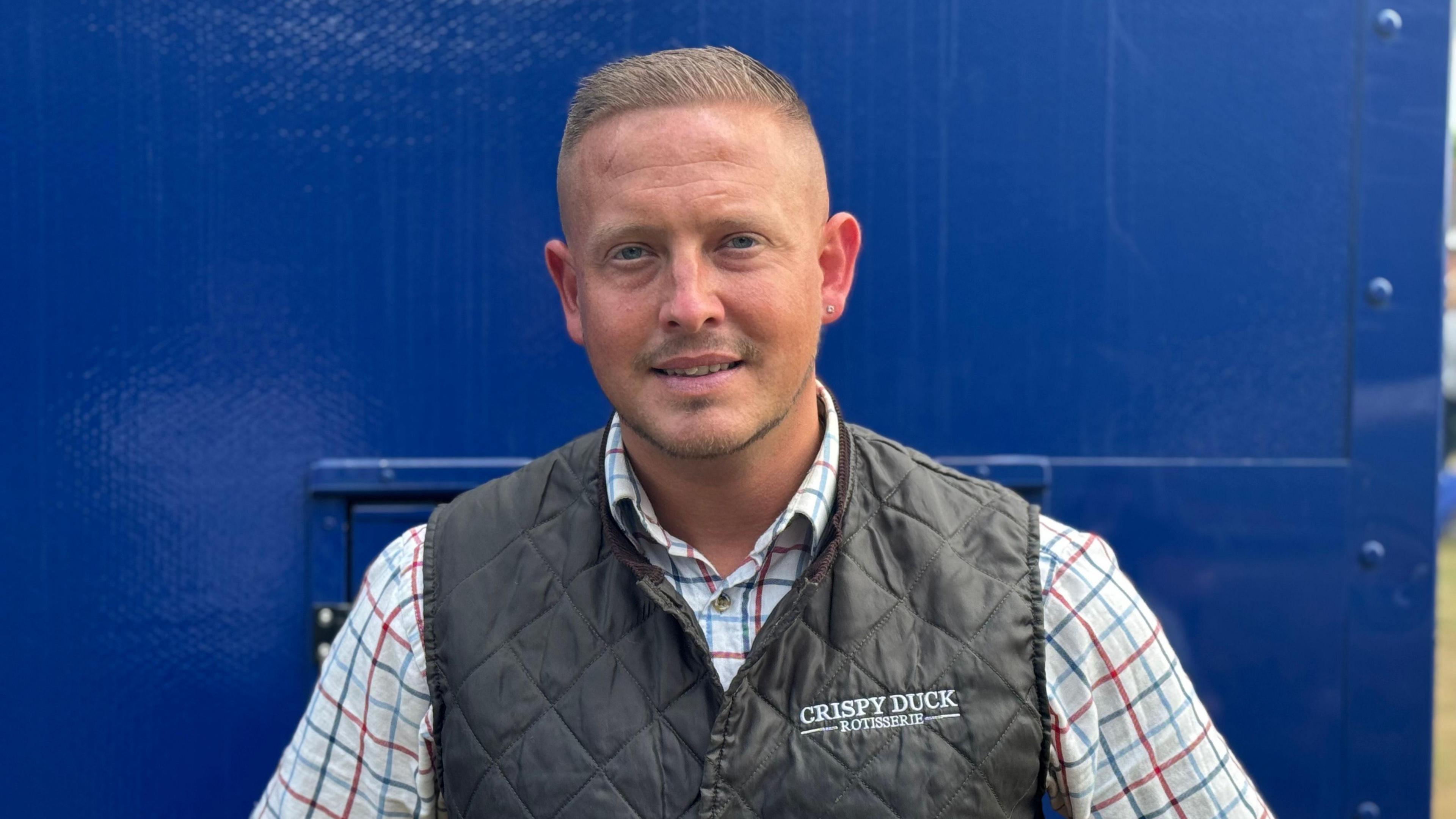A head and shoulders shot of Shaun Sabiny, who is standing in front of a navy blur food trailer. He is wearing a gilet over a shirt and is smiling into the camera.