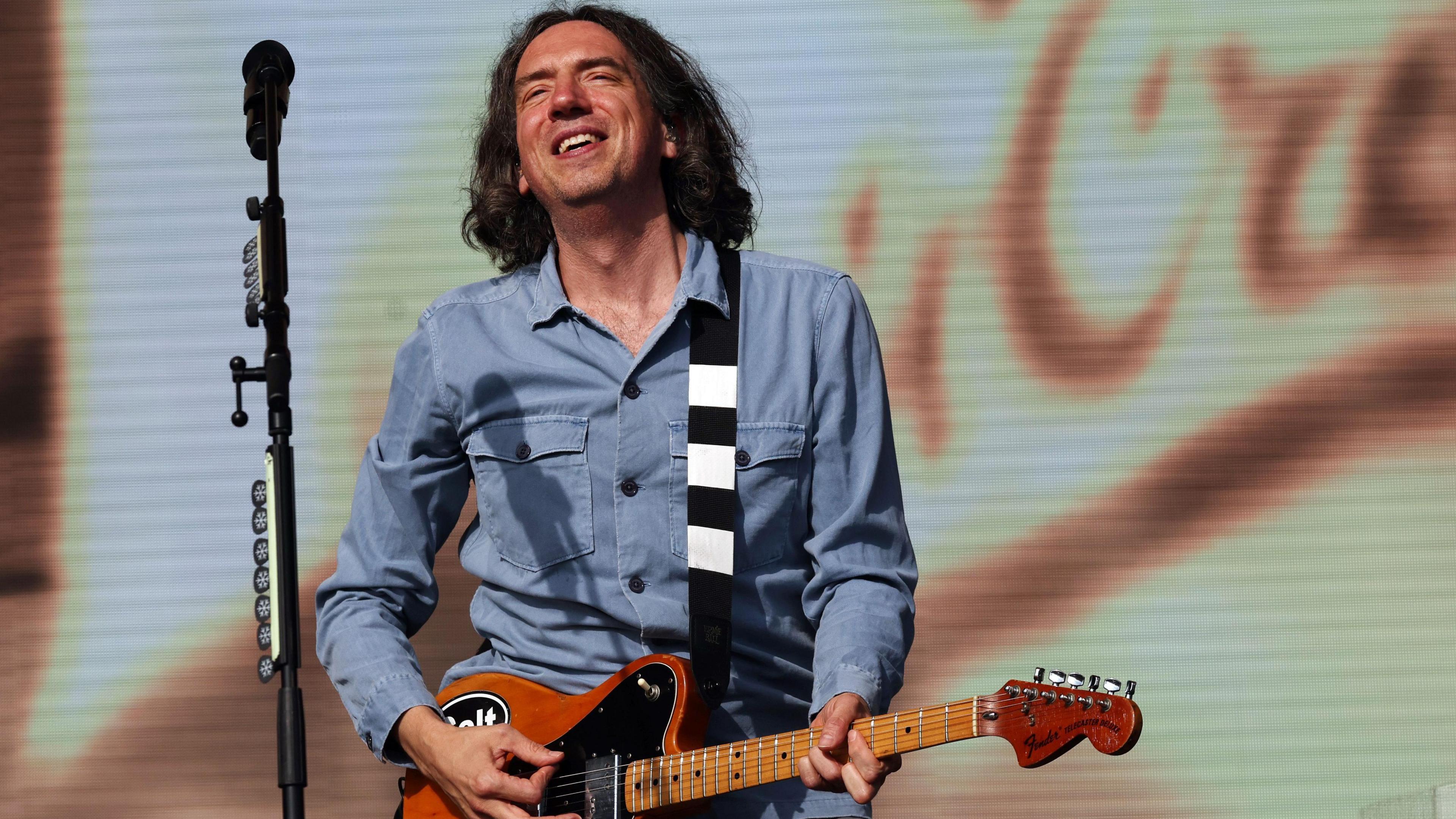 A man in a blue shirt is holding an electric guitar on a stage in front of a microphone stand.