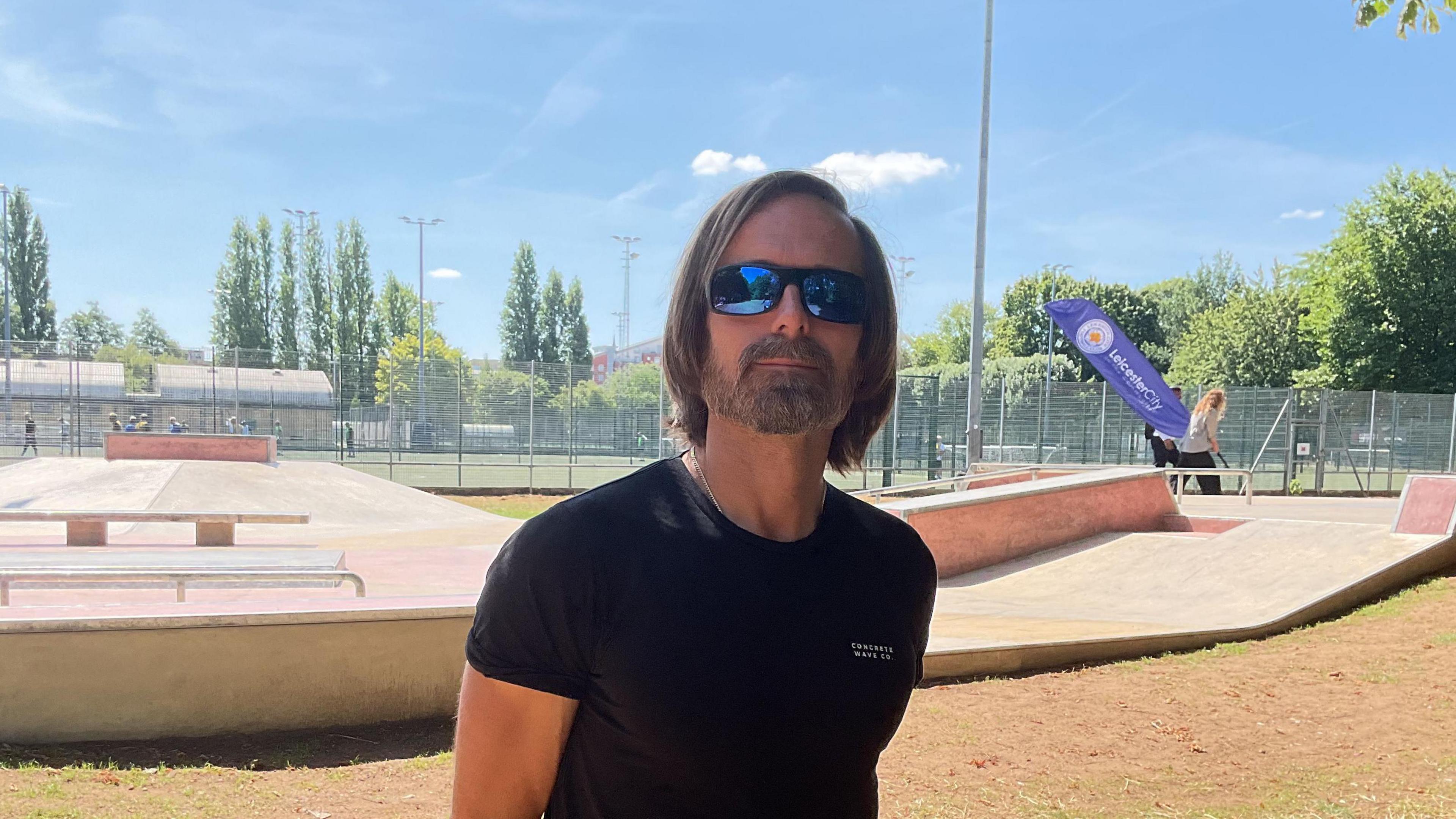 Russ Holbert wearing a black T-shirt and sunglasses in front of the skatepark