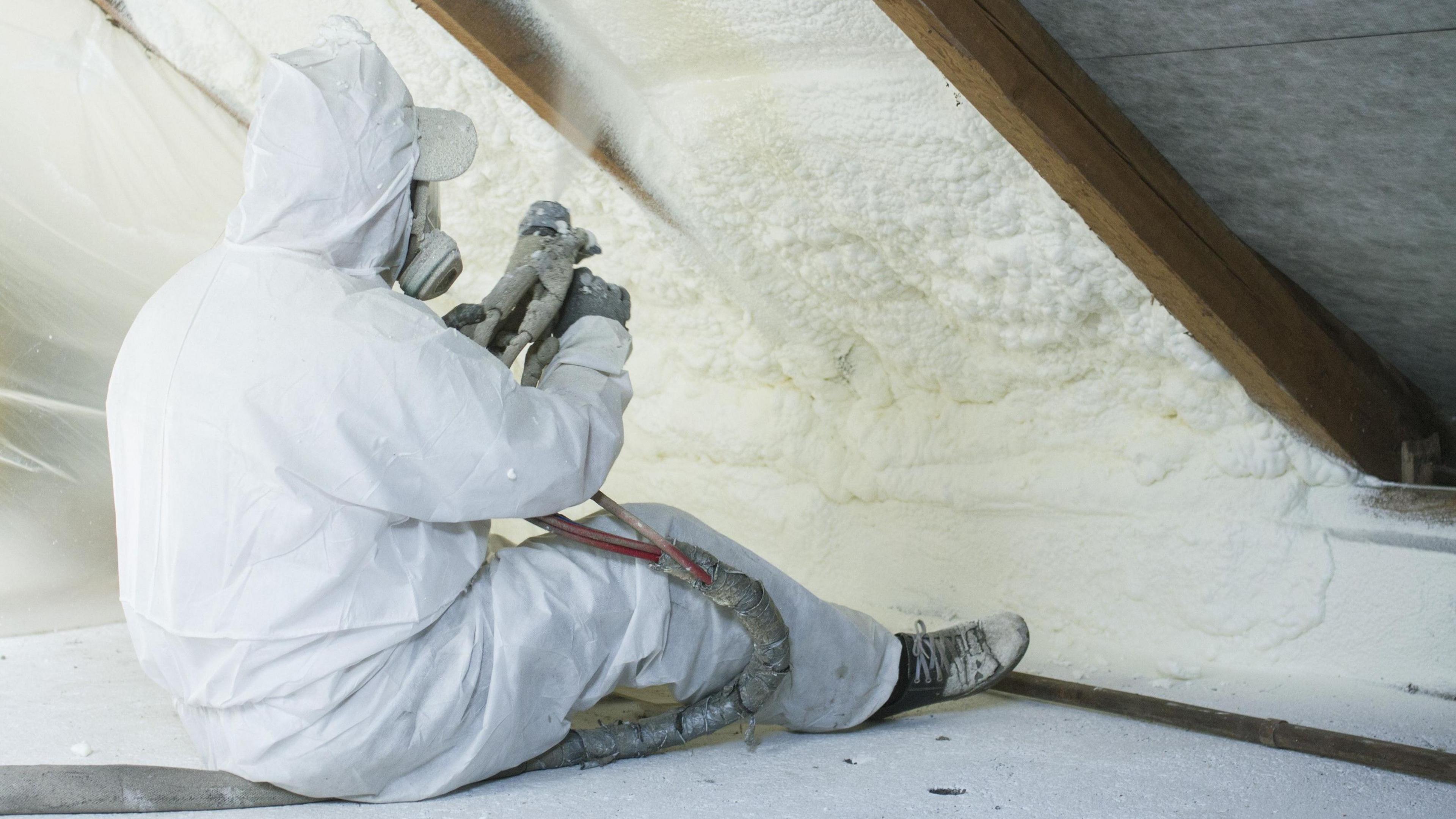 Roof technician spraying foam insulation using component gun while wearing a white boilersuit