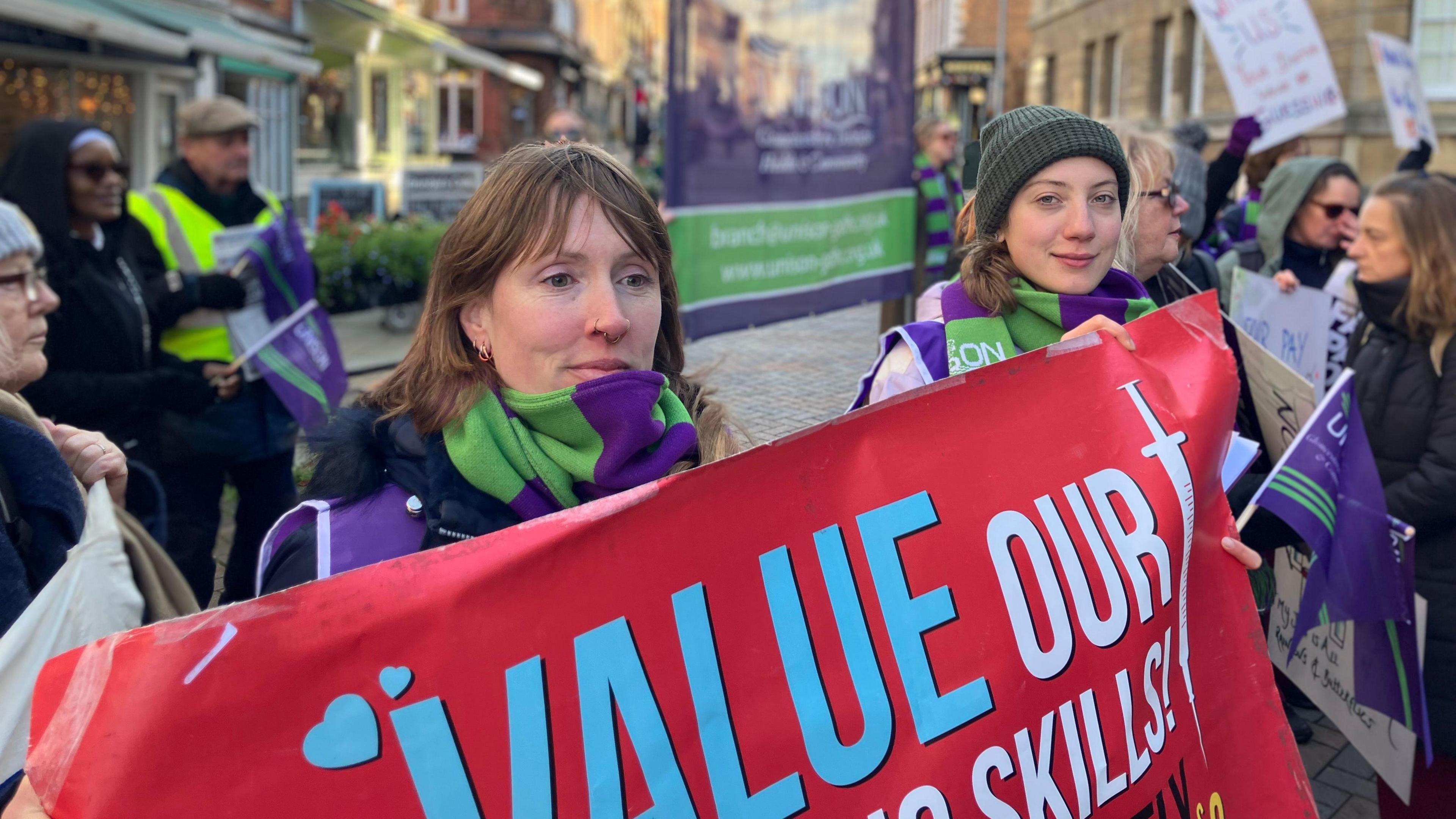 A woman with light brown hair to her shoulders holds up a red banner at a protest, along with another woman wearing a beanie. They are bundled up for the cold weather in green and purple scarves. Other people at the rally can be seen around them, holding flags and placards on a city centre, pedestrianised street.