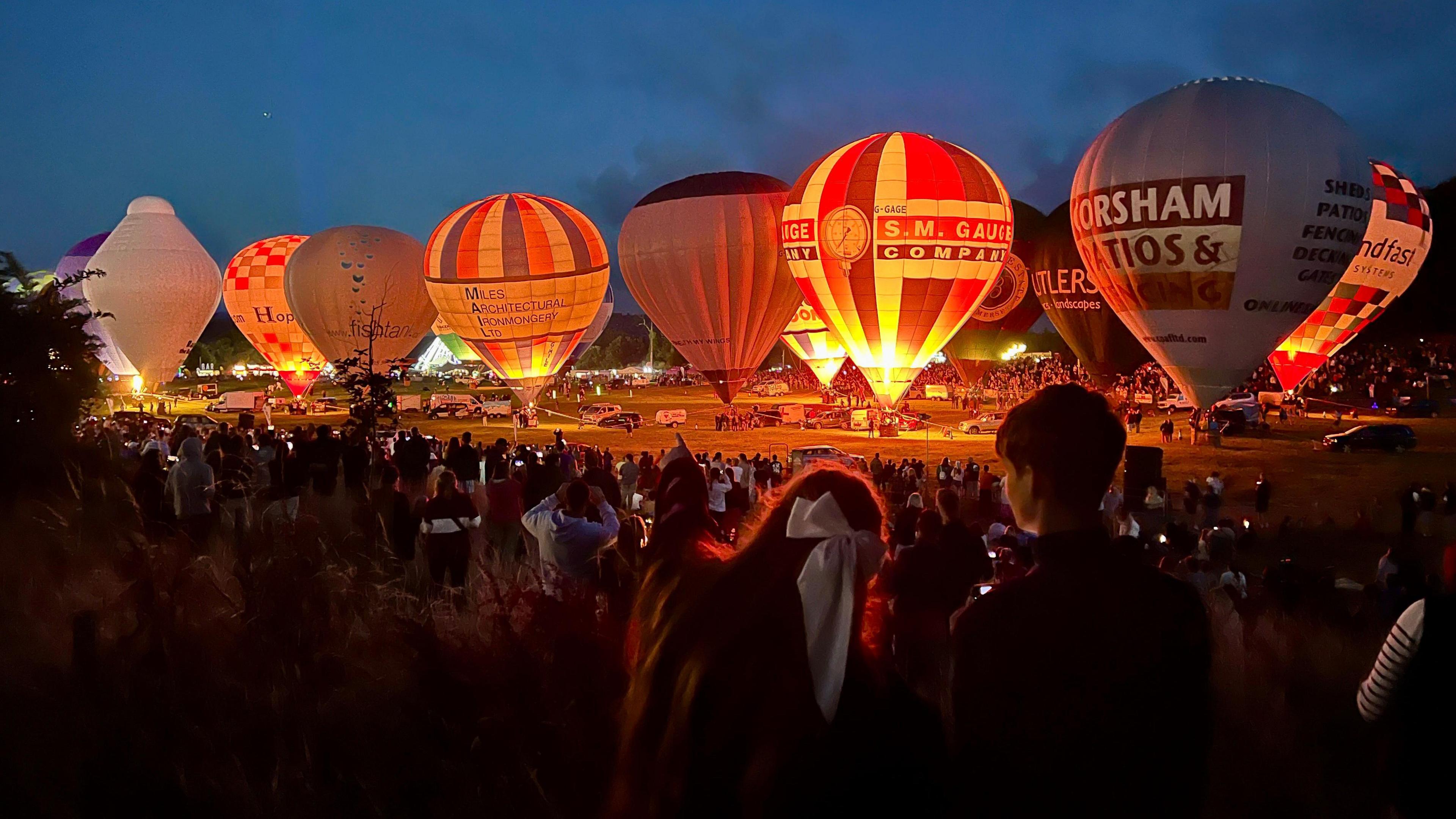 A crowd of people are seen looking and pointing at a row of inflated hot air balloons that are lit up in the dark.