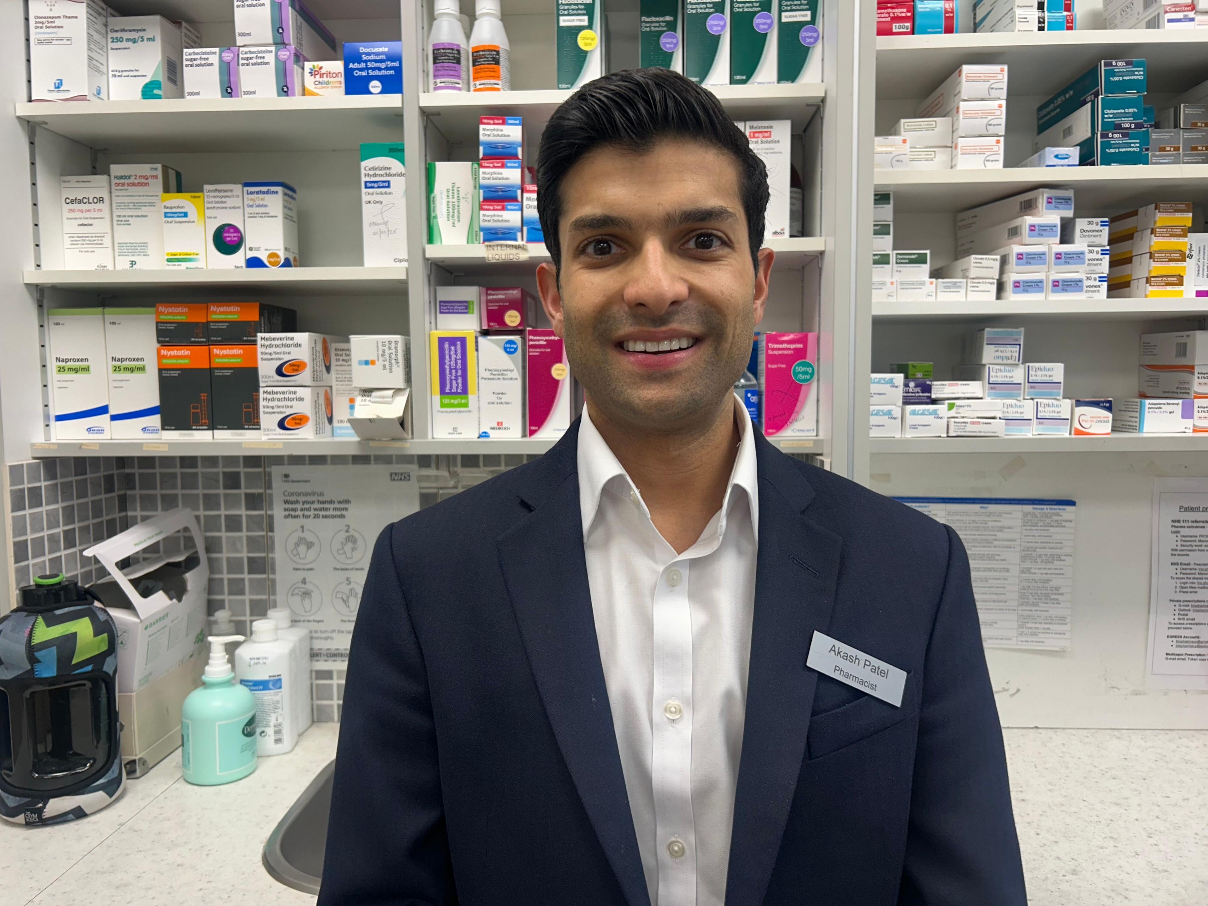 Një i ri me këmishë të bardhë dhe xhaketë blu të errët qëndron në një farmaci duke i buzëqeshur kamerës.