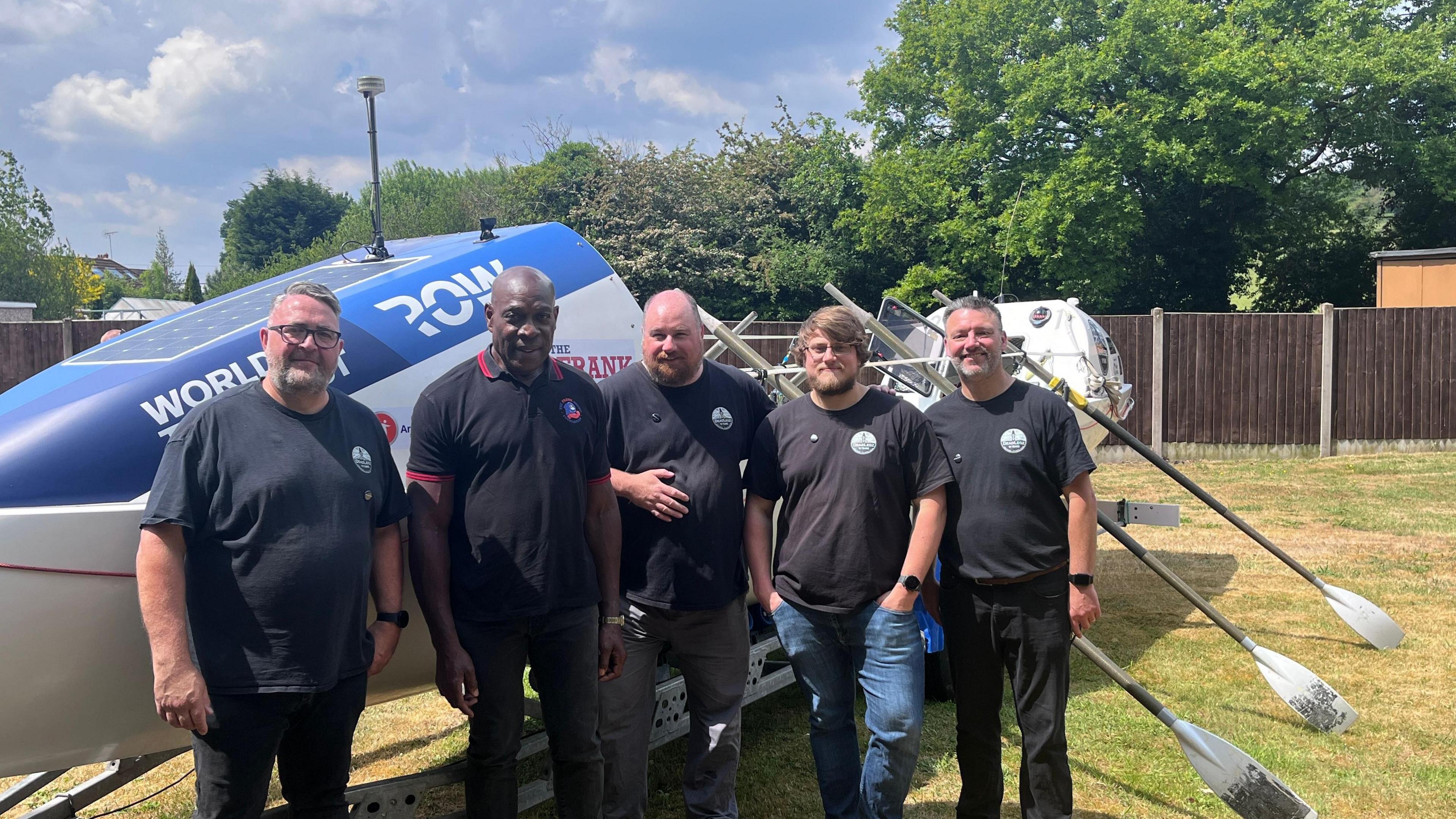 Frank Bruno stands with the group that are rowing the Atlantic, with the boat behind them and oars to one side. Left to right, the men seen in the picture are Rob Oliver, Matt Humphries, Tom Hayward and Kev Joynes.