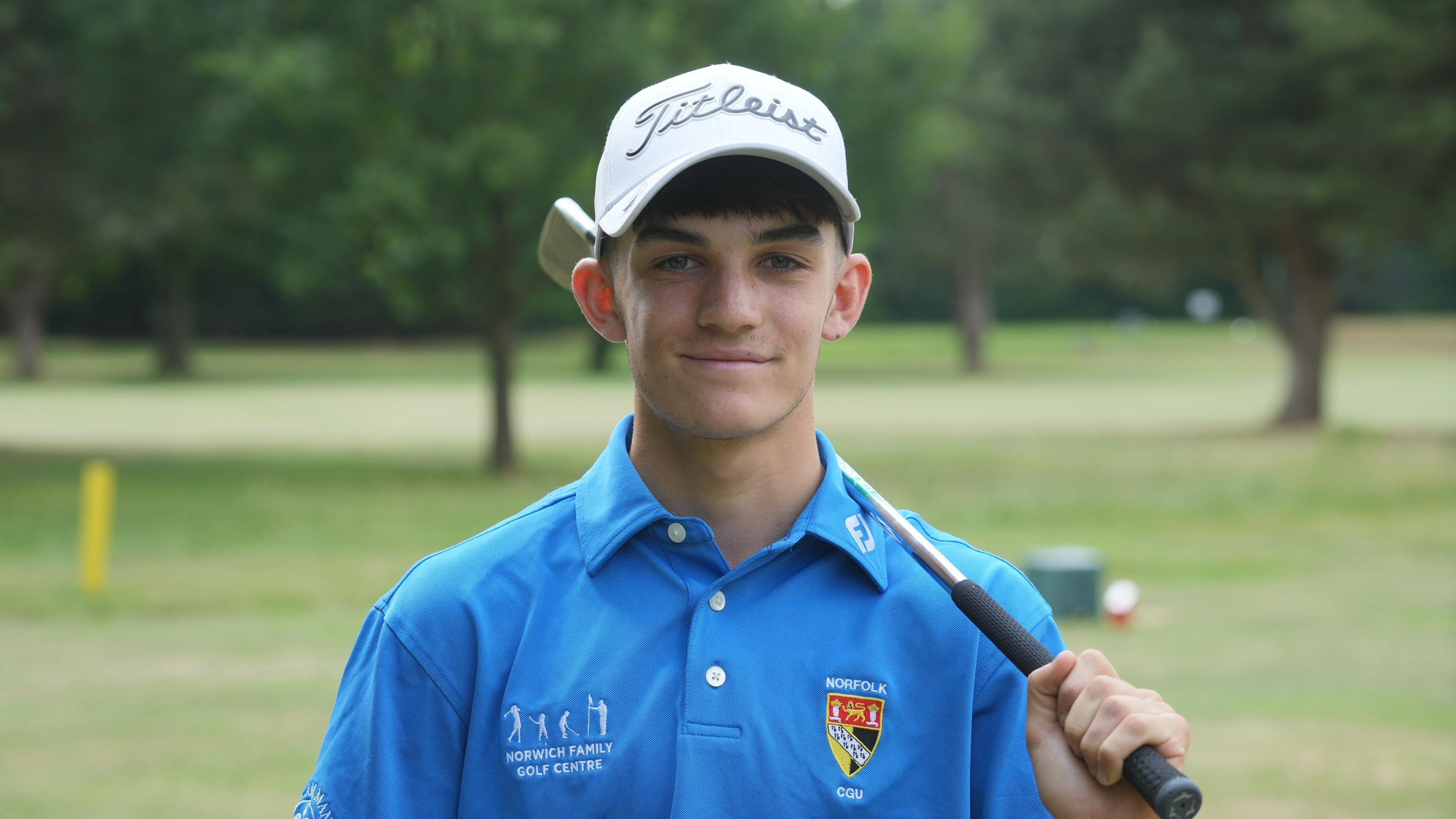 Liam Caffyn poses for a photograph in a blue Norfolk County Golf Union shirt with a club draped over his left shoulder. There is a golf course with trees in the background.