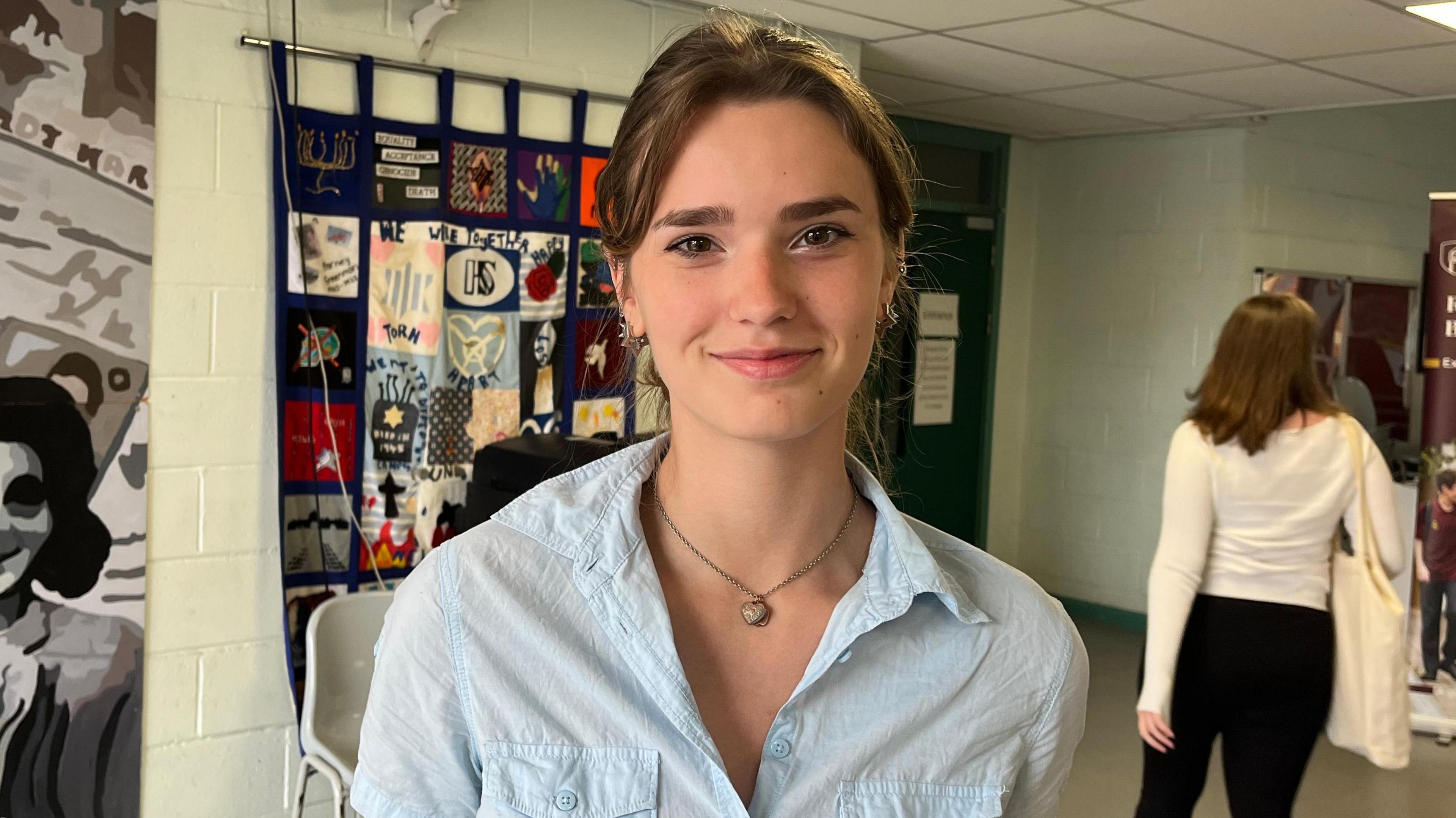 Taylor smiles at the camera. She has brown hair, which is tied back, and wears a pale blue shirt and a short necklace with a heart shaped locket on it. 