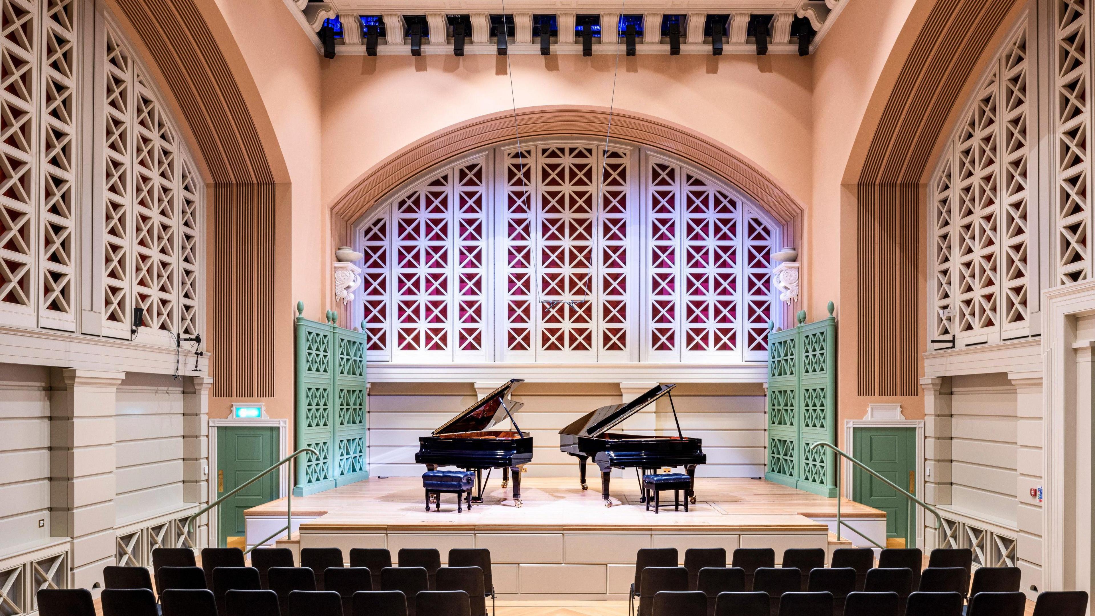 A hall with chairs lined in rows and on stage are two black grand pianos. There is green panelling on either side of the stage with two green doors on either side of the stage. 