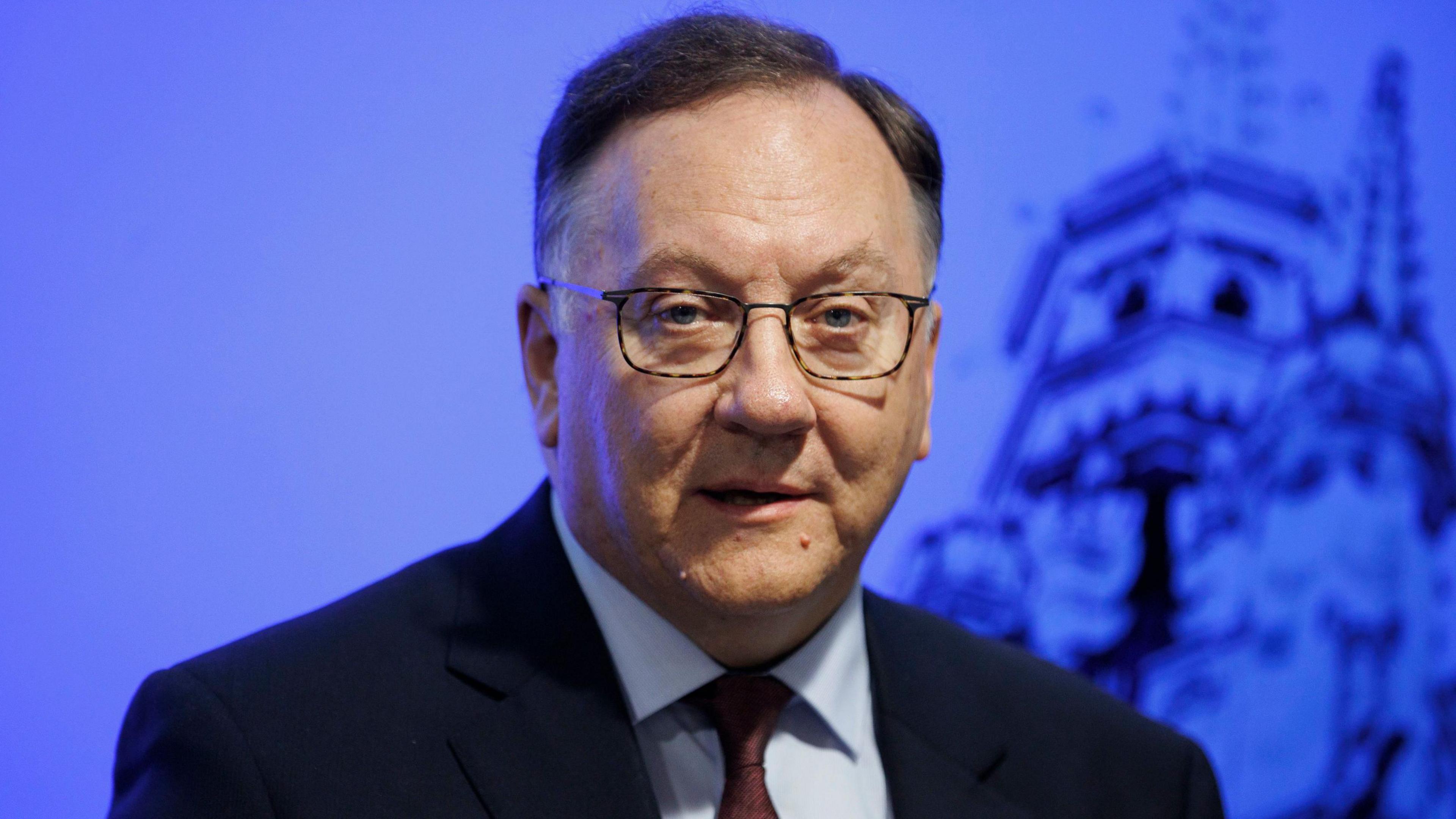 Rev Trevor Gribben, pictured from the chest up. He is wearing a black suit, blue shirt and dark red tie. He has short brown hair. He is standing in front of a blue background.