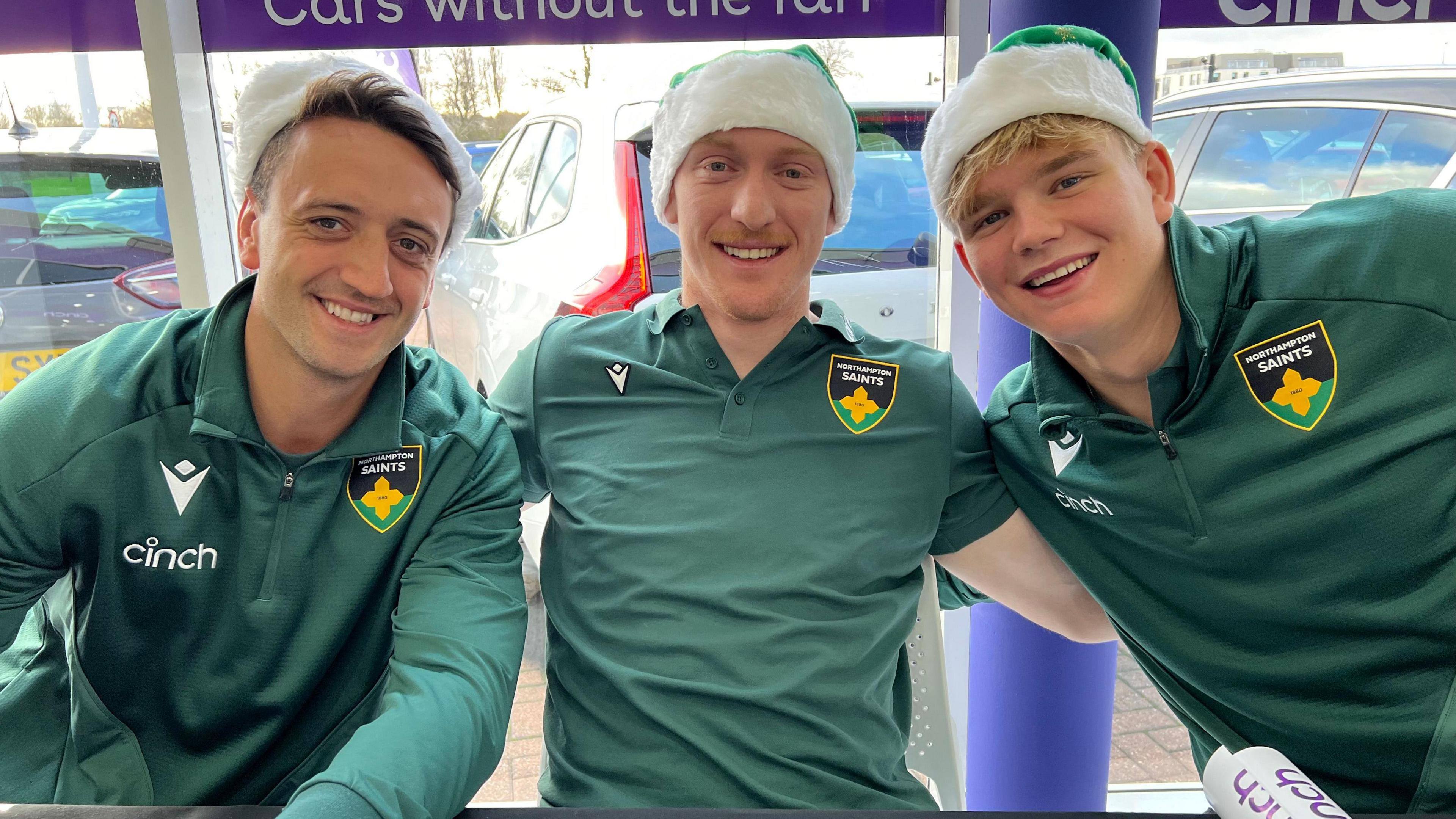 Alex Mitchell, George Hendy and Henry Pollock sit side by side. All three are wearing green Saints training tops and green "Santa" hats. All three are looking at the camera and smiling.