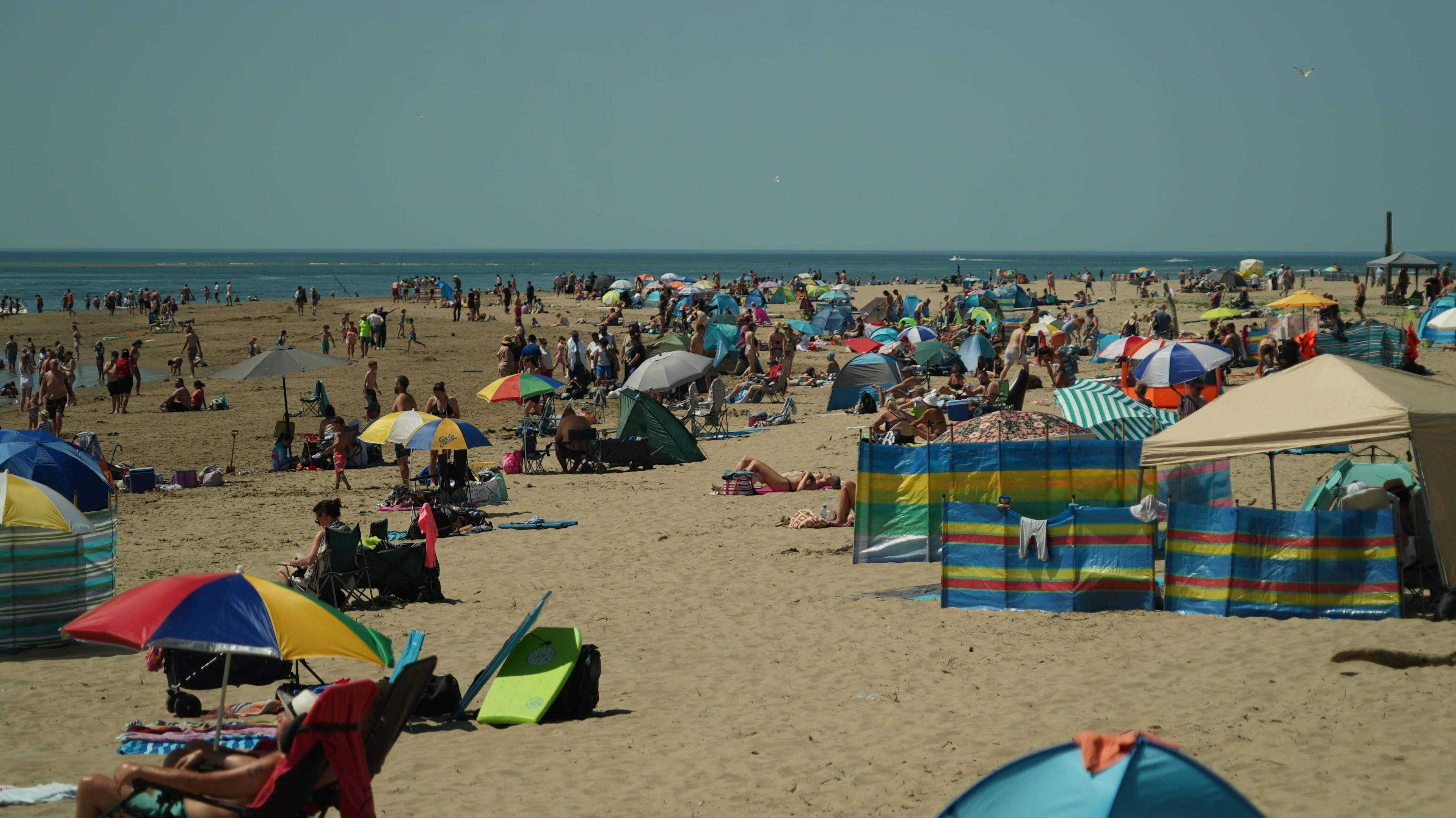 A packed beach filled with sunbathers and parasols and the sea in the background.