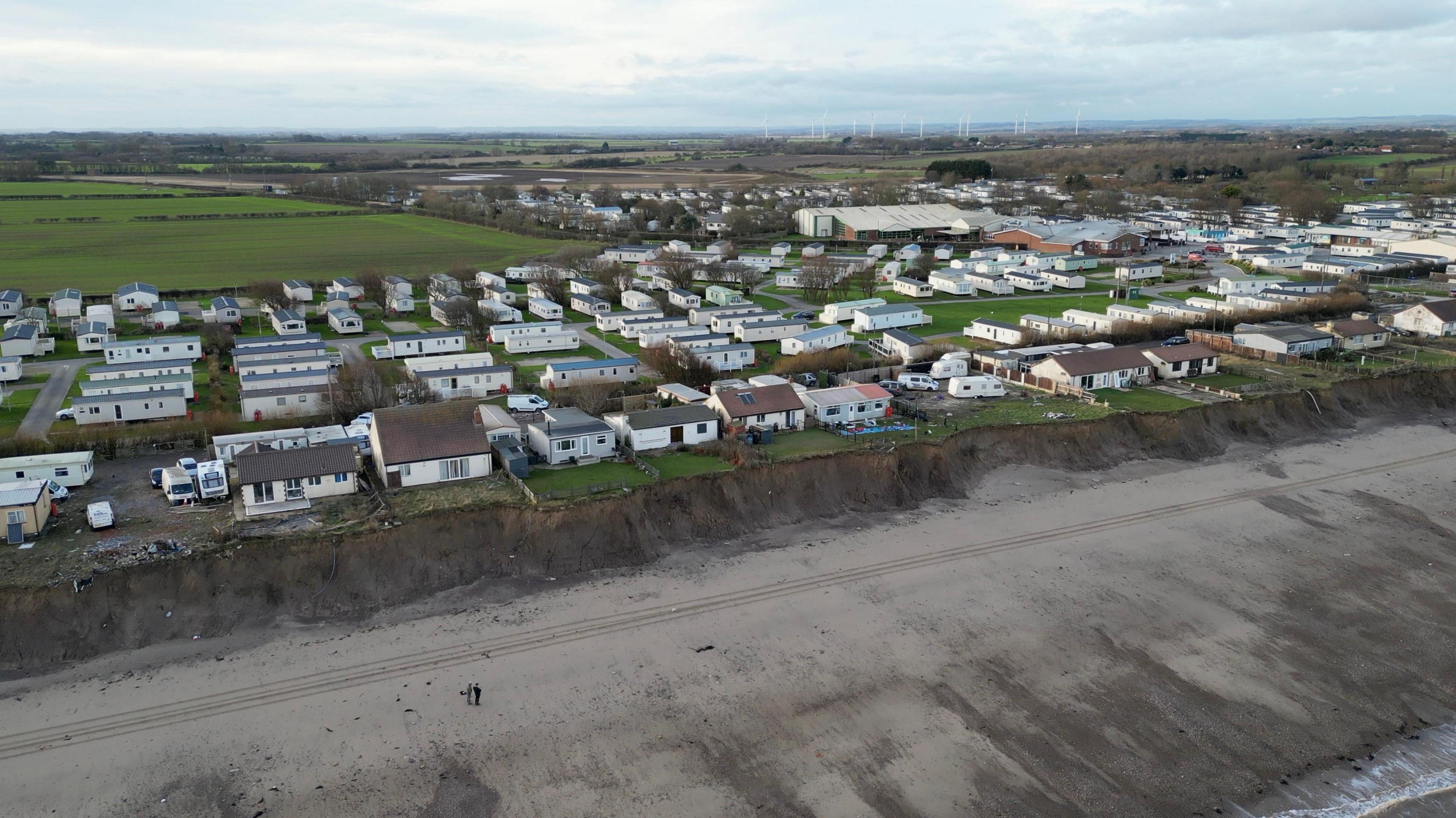 An aerial view of Skipsea captured by a drone on the beach. The sand and sea are visible in the lower part of the image which then leads to a muddly looking cliff edge. A number of houses and static caravans are visible on the clifftop and, in the distance, fields and wind turbines can also be seen against the cloudy sky.