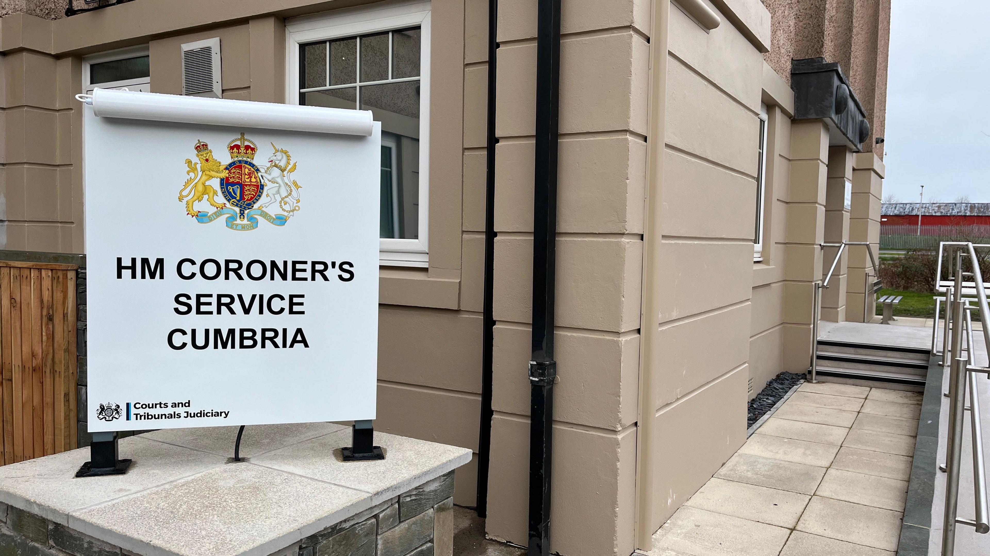 A sign outside the coroner's court in Workington says "HM Coroner's Service Cumbria". Next to it is a ramp leading to the court's entrance. The building is painted in a hazelnut colour.