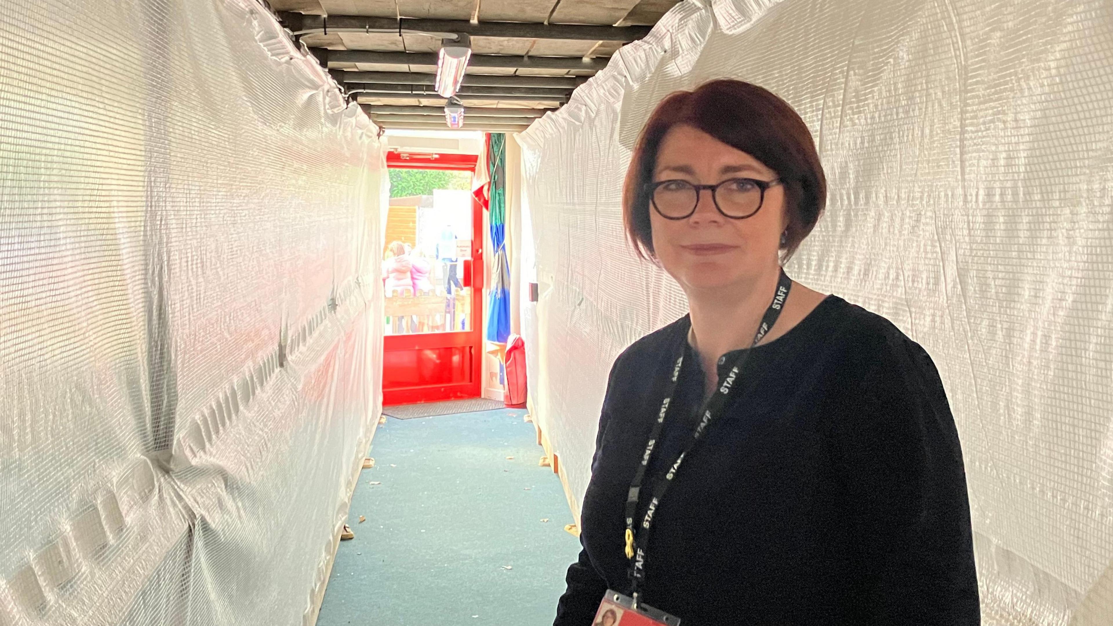 A head-and-shoulders shot of headteacher Becky Black, who has a dark hair in a bob-style cut and is wearing glasses and a dark, long-sleeved top. She has a school lanyard around her neck. She is standing in a school corridor. The walls on each side of the corridor are covered by builders' plastic sheeting. There is a red door at the end of the corridor.