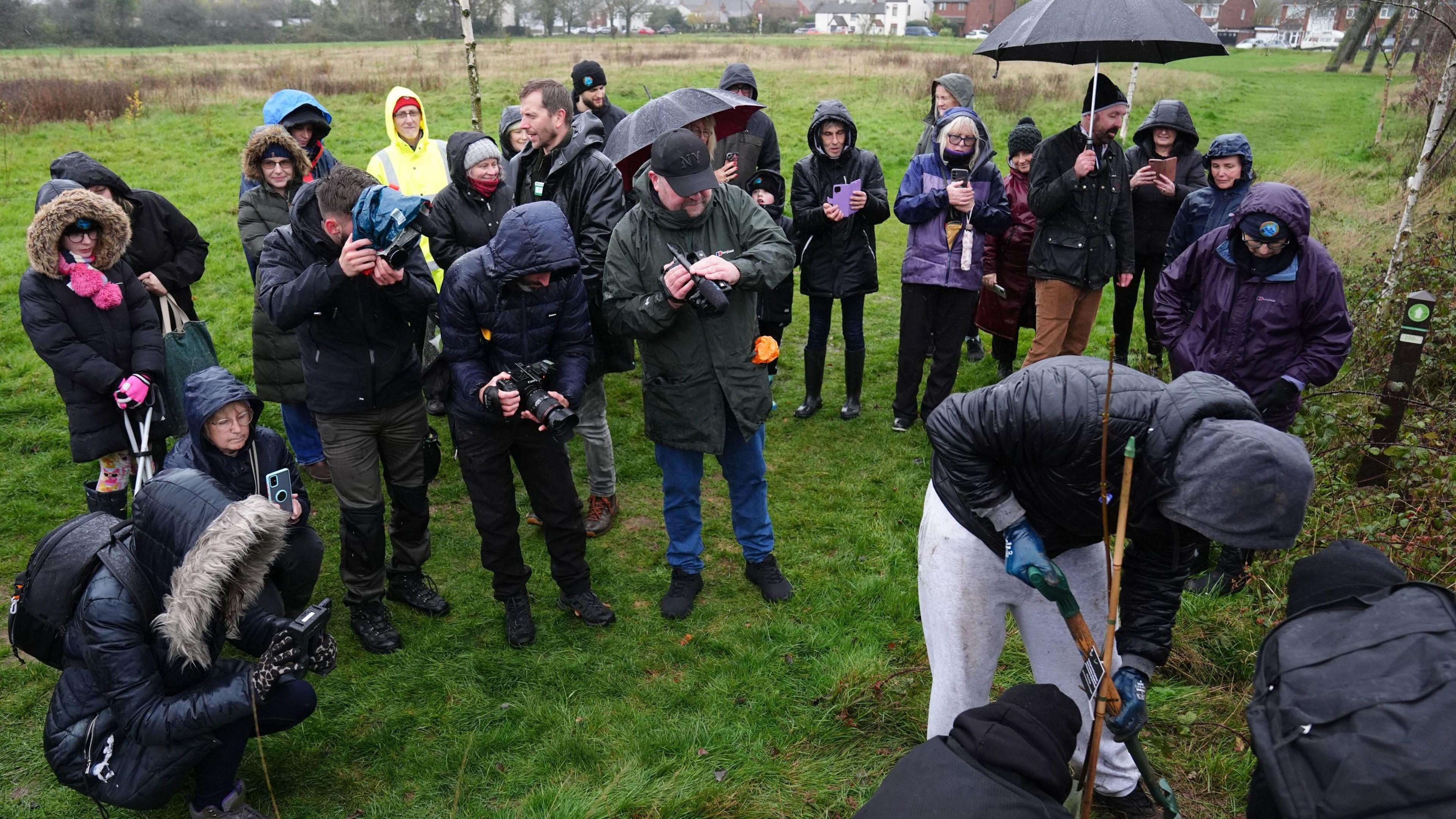 Thomas Irwin from The Tree Amigos volunteer group plants a Tree of Hope at the Tree Sanctuary in Coventry. People are gathered round to watch. They are wearing raincoats and carrying umbrellas on a rainy day. A woman is crouching and taking a photograph.