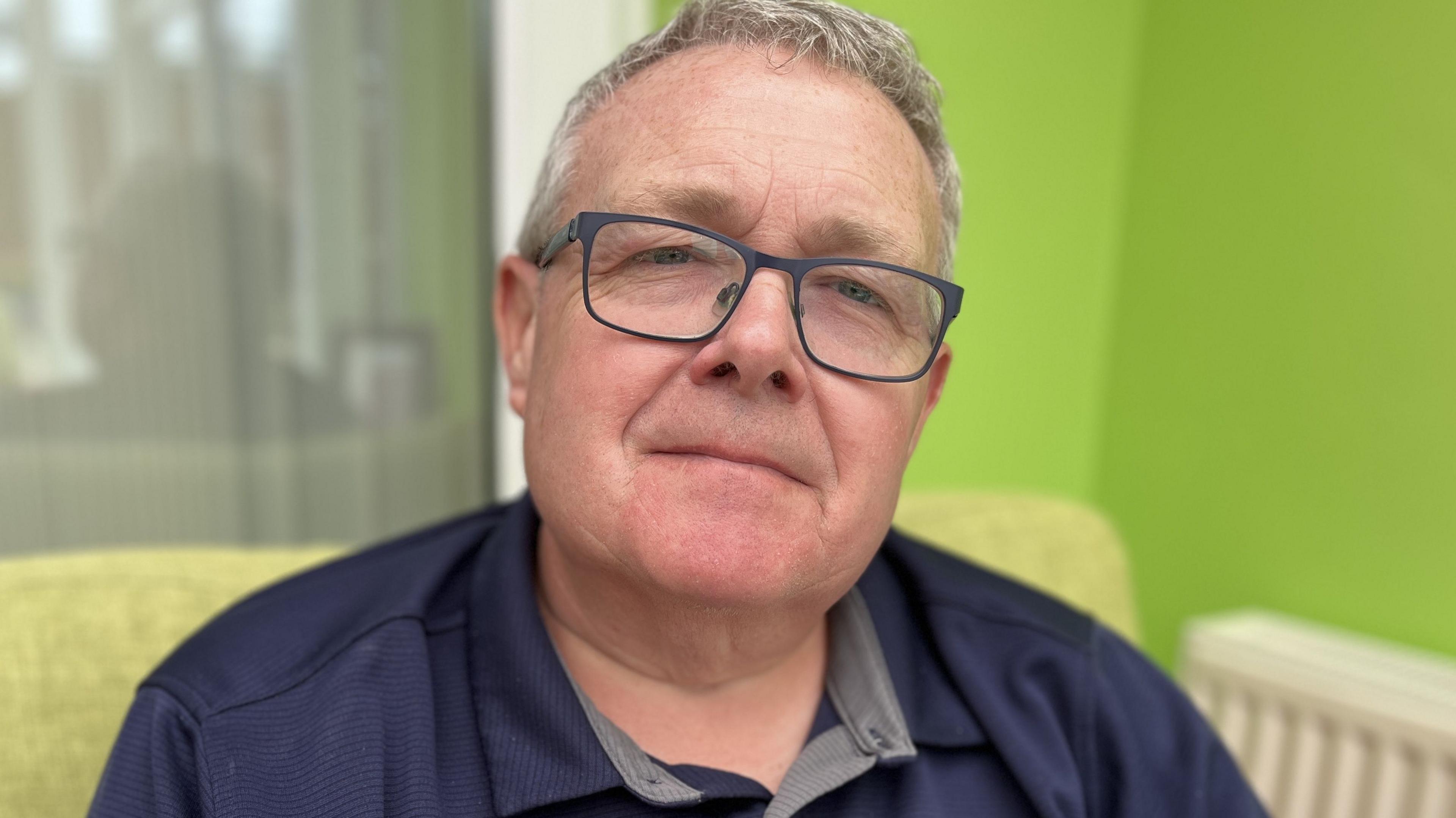 Steve Blanchard who is wearing black rimmed glasses and short grey hair. He is sat down and there is a lime green wall with a radiator on behind him. 