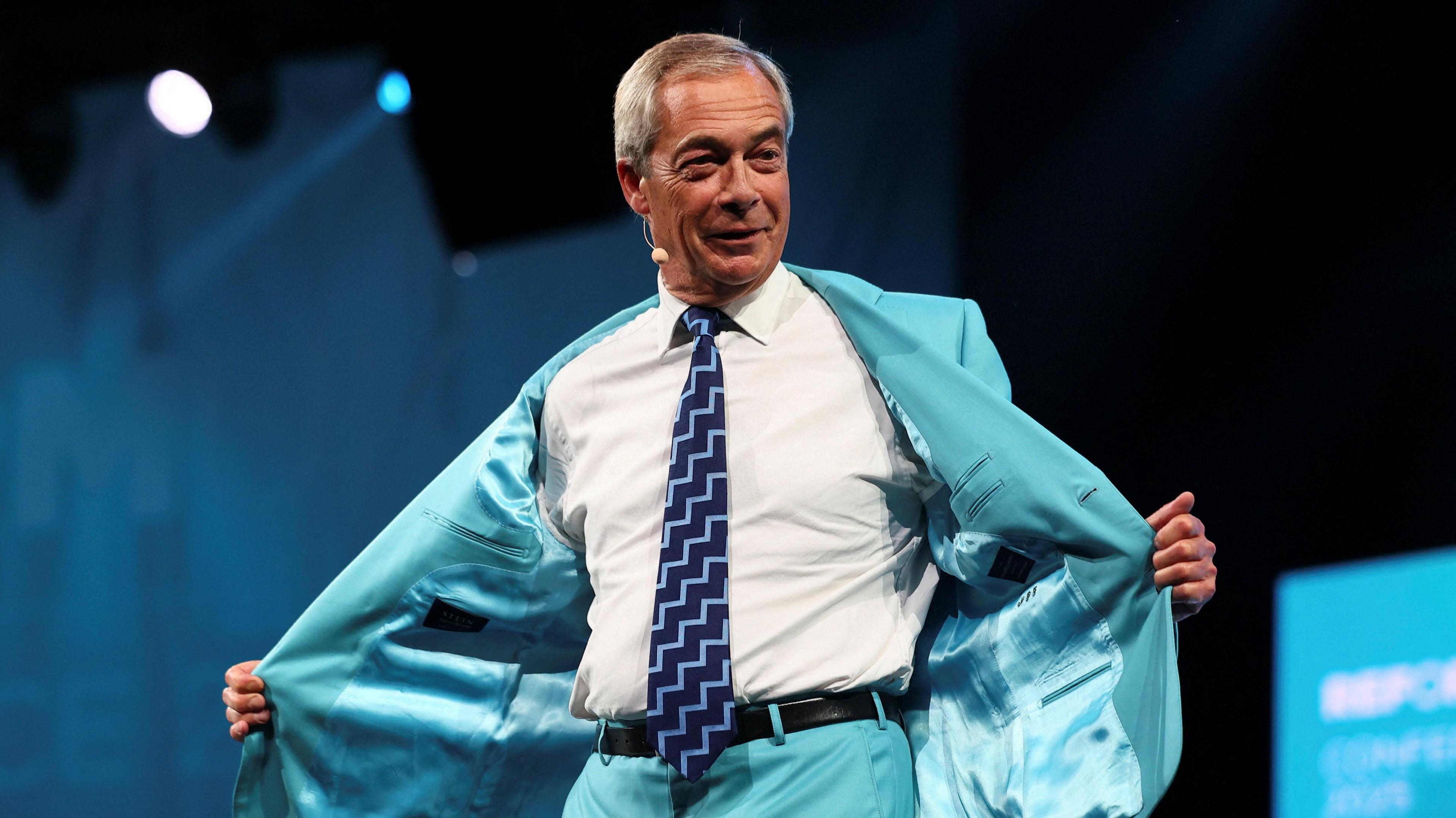 Nigel Farage has short grey hair and is wearing a white shirt, blue tie and turquoise suit. He is holding open the suit jacket, which reveals a satin-type lining.