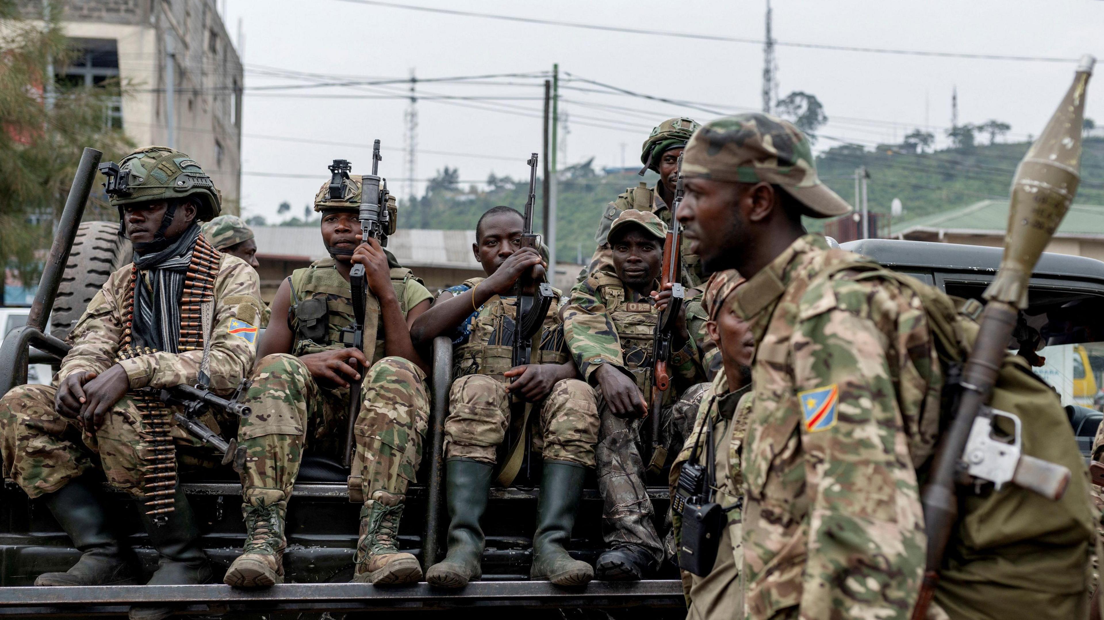 Four armed militants wearing camouflage and holding guns sit on the back of a truck as another soldier with a grenade launcher on his shoulder walks past in the foreground