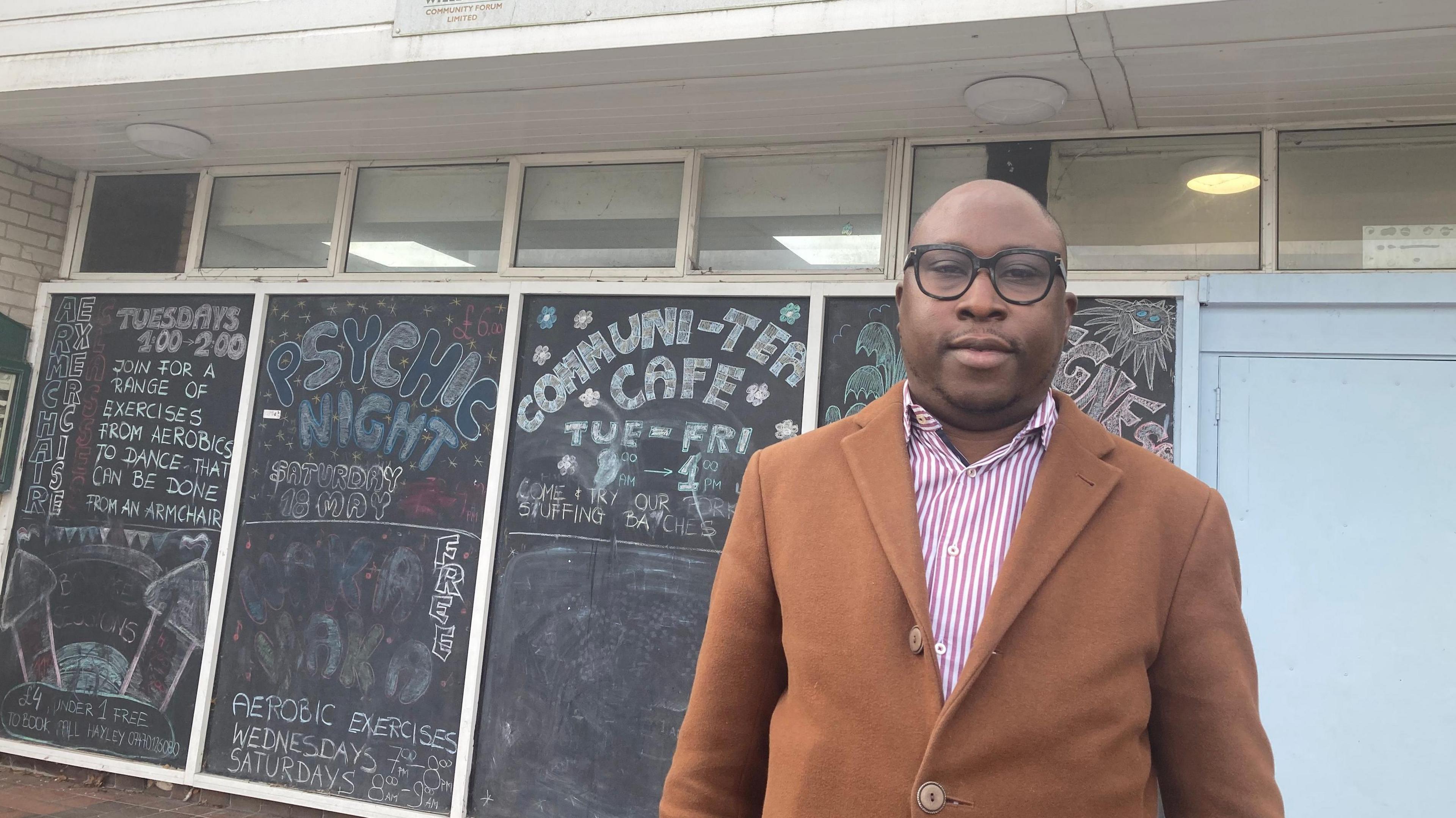 A man with round, black-rimmed glasses, a smart tan-coloured coat over a pink and white striped shirt. He is standing in front of the community centre and there are blackboards with chalk written over them, describing all the community events that take place there.