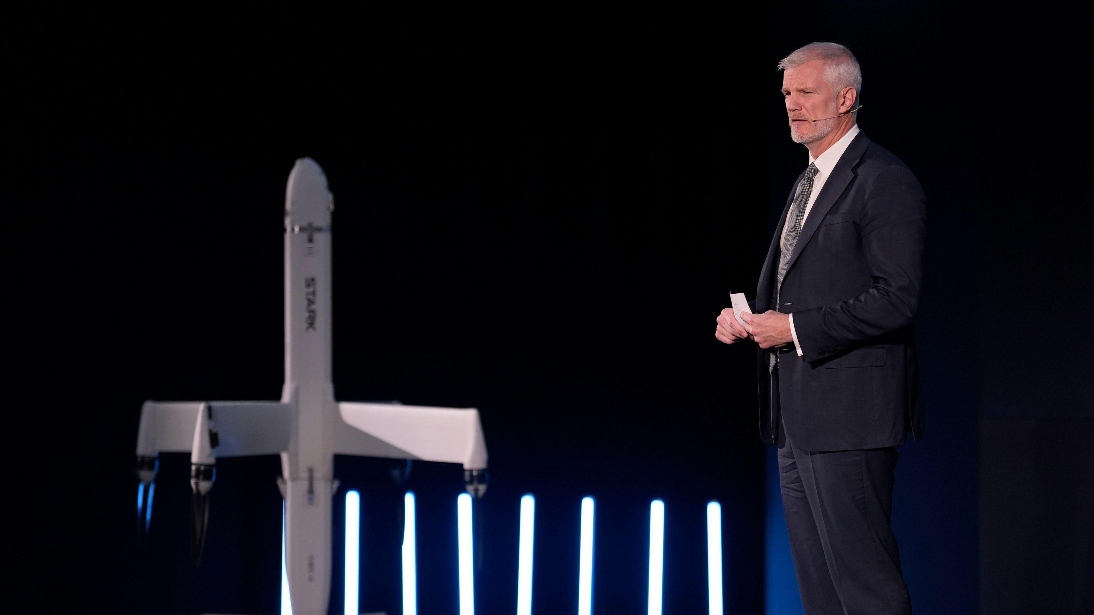 Alistair Carn standing on a stage giving a speech in a suit and tie. He has a model of a drone next to him
