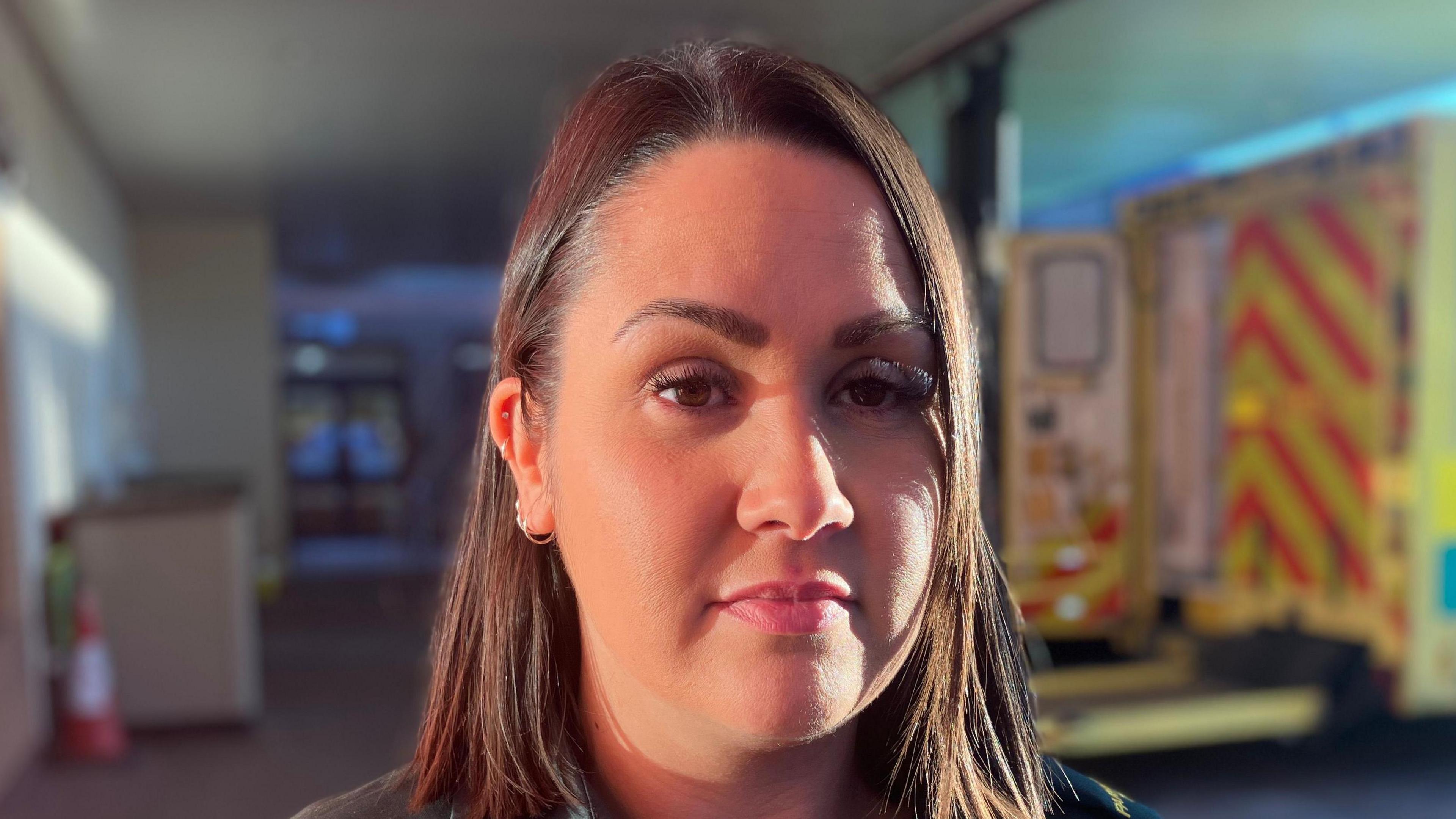 Chloe Lancey has shoulder-length brown hair and is wearing a paramedic's uniform and standing in front of an ambulance 