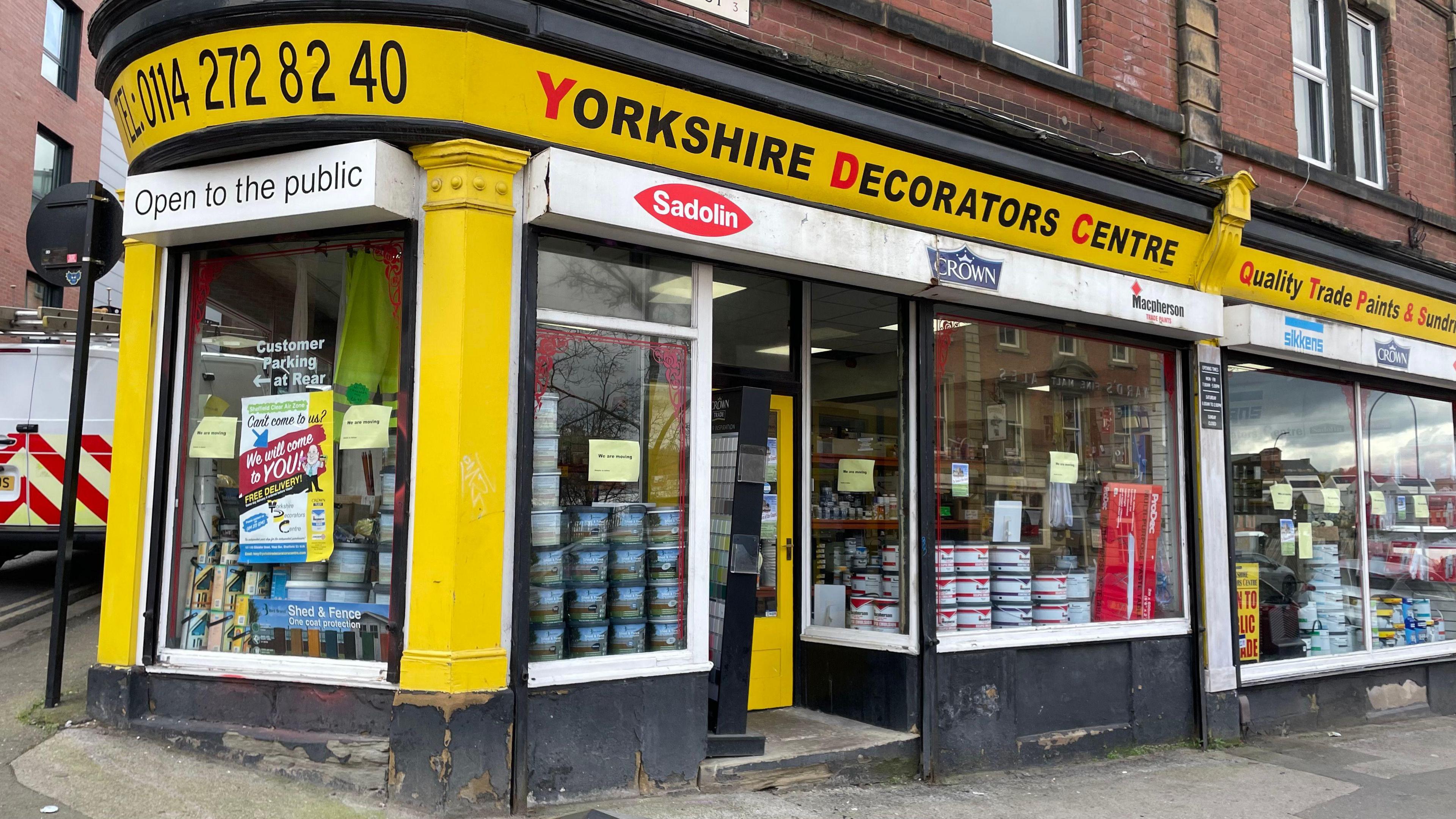 The front of the shop, which has been painted yellow, and has stacked tins of paint in the windows.