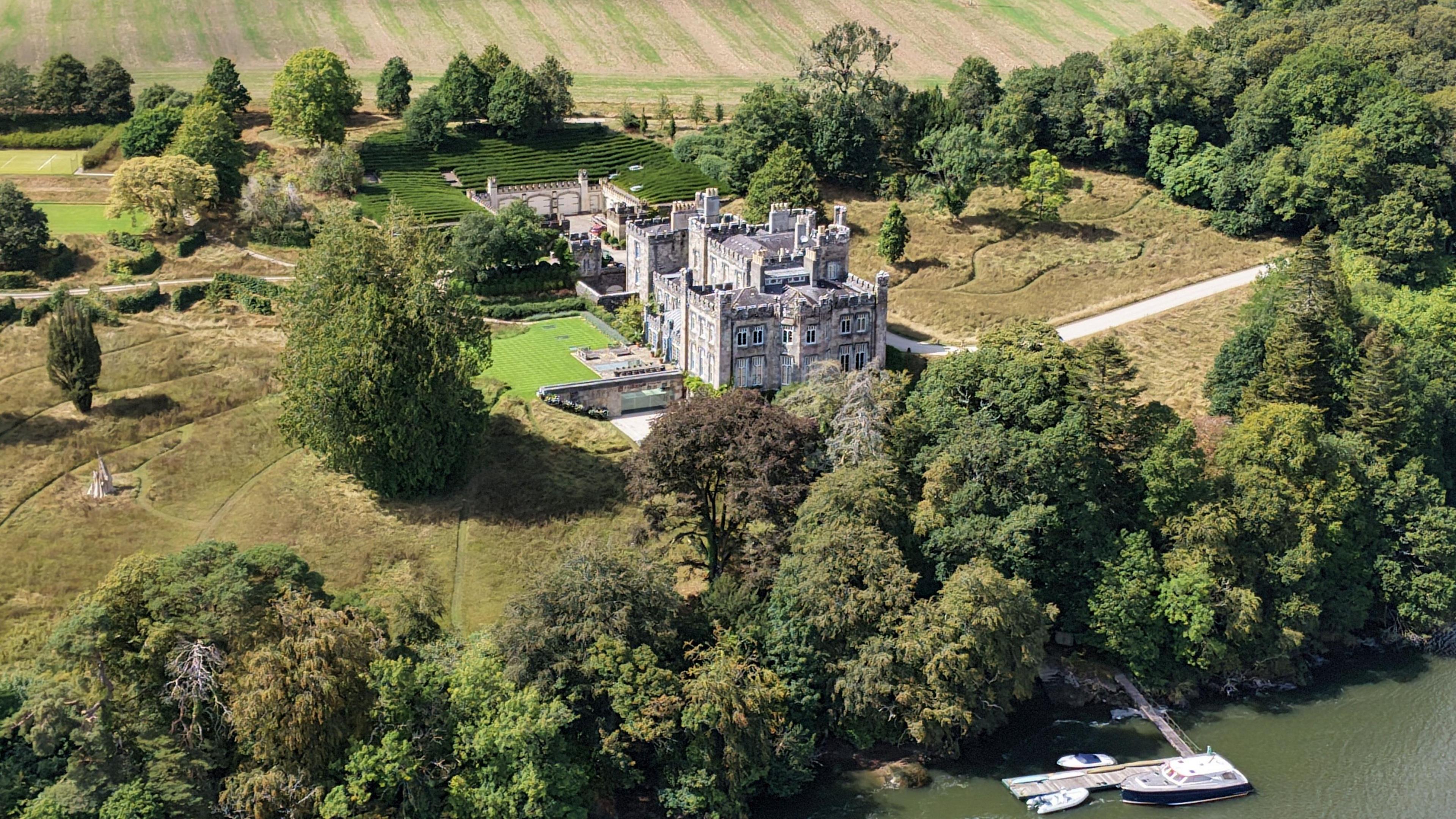 An aerial view of a castle, surrounded by trees and fields. It is overlooking a river.