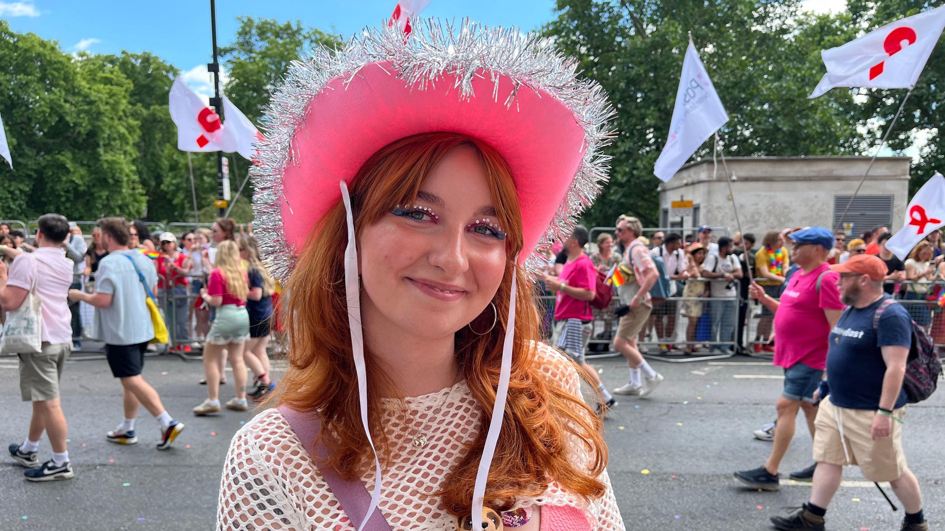 A medium close-up of a smiling attendee named Magda Szewczak, who has red hair and glitter eye makeup, wearing a bright pink cowboy hat trimmed with silver tinsel at the parade.