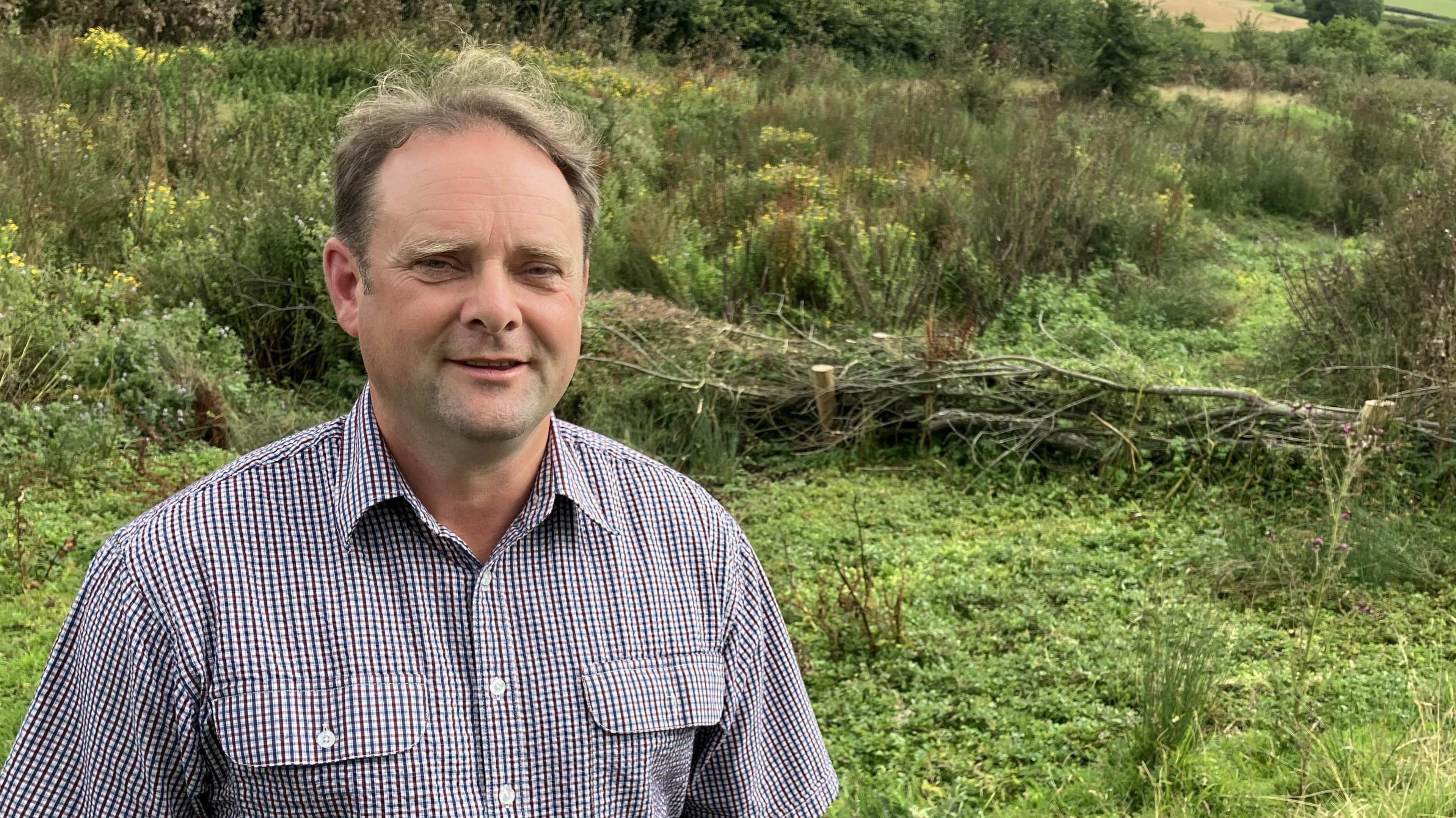 A man in checked shirt stands in a field with a small dam across a river in the background