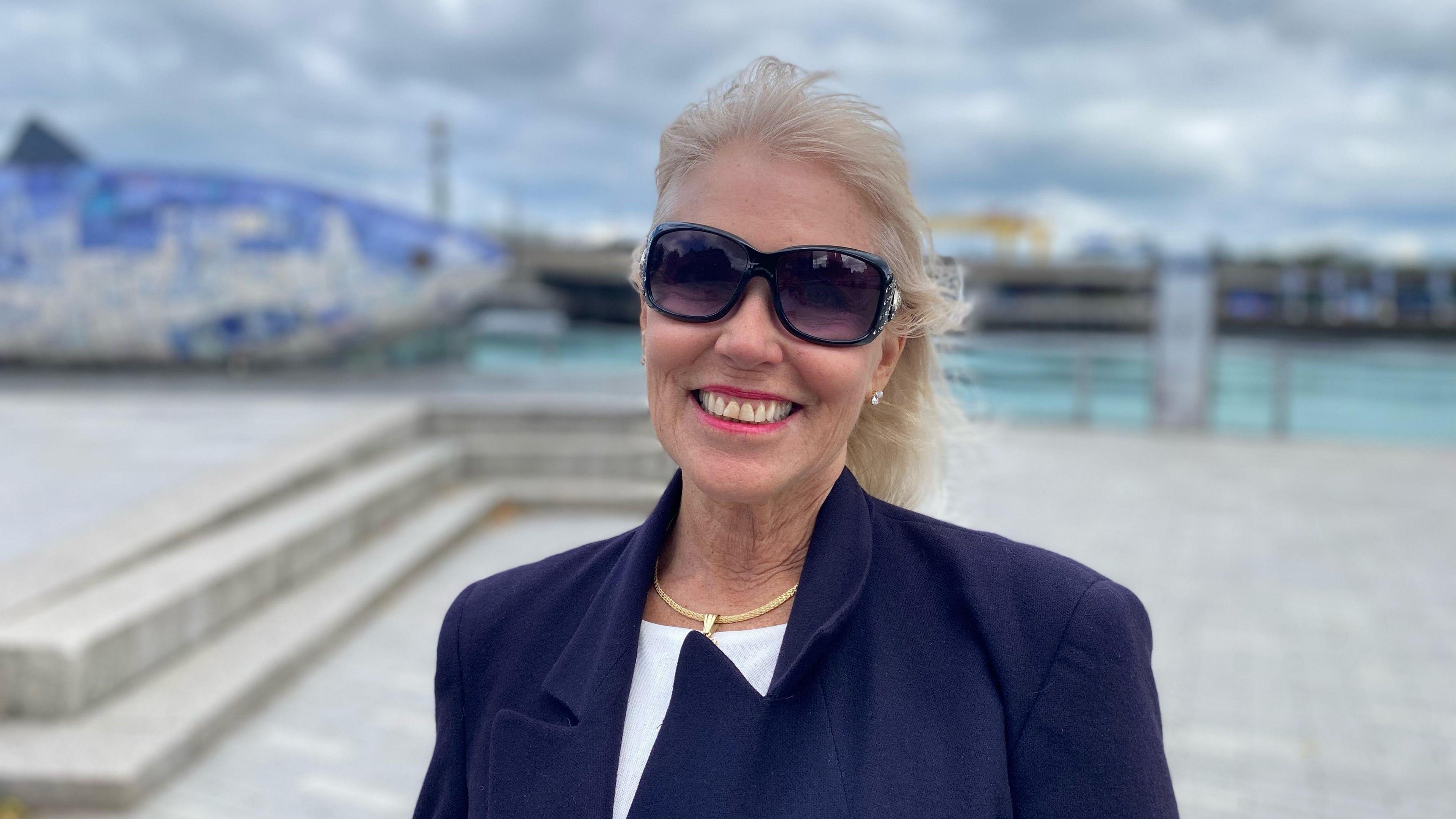 Blonde haired woman, windswept wearing large sun glasses and a navy blazer.