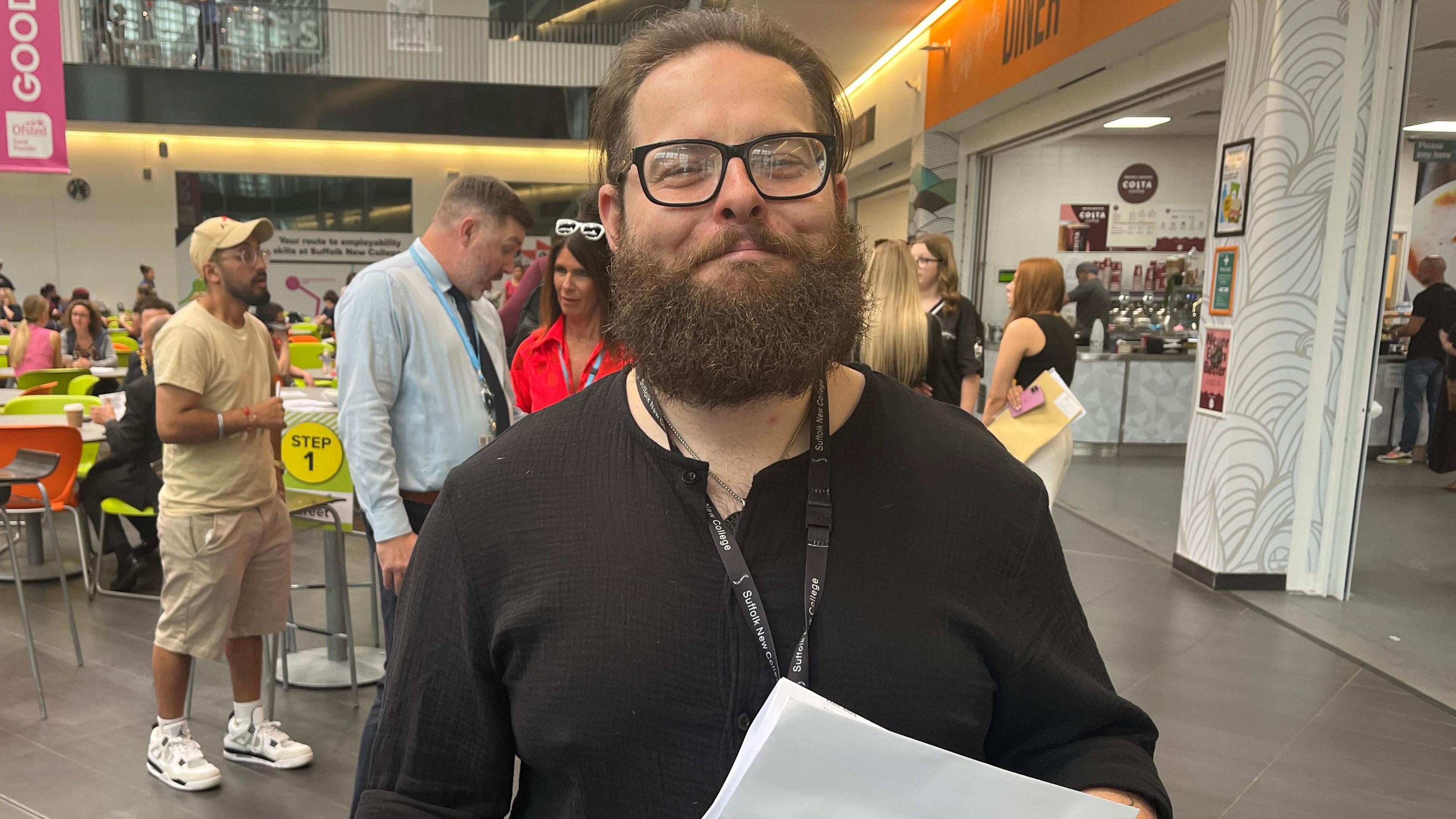 James Brown is standing in the middle of the image with a slight smile. He has short brown hair and a long brown beard, and is wearing a black T-shirt and holding a white piece of paper.