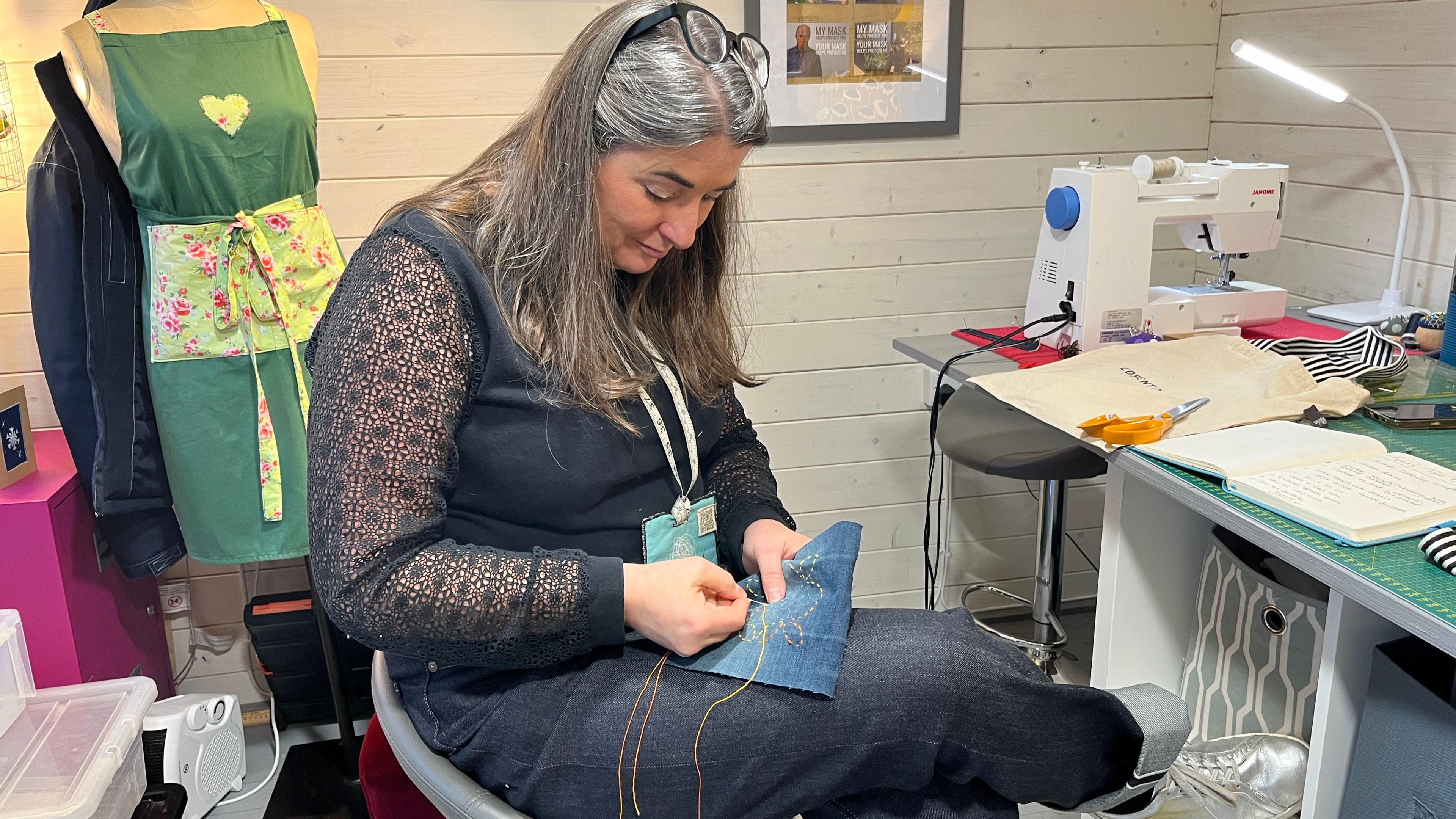 Melissa Santiago-Val is sat in a sewing room. She is hunched over as she looks at a pattern she is sewing with ha needle and thread. She has long brown hair, a blue blouse and blue jeans on. In front of her is a crafts table with sewing equipment and a sewing machine. 