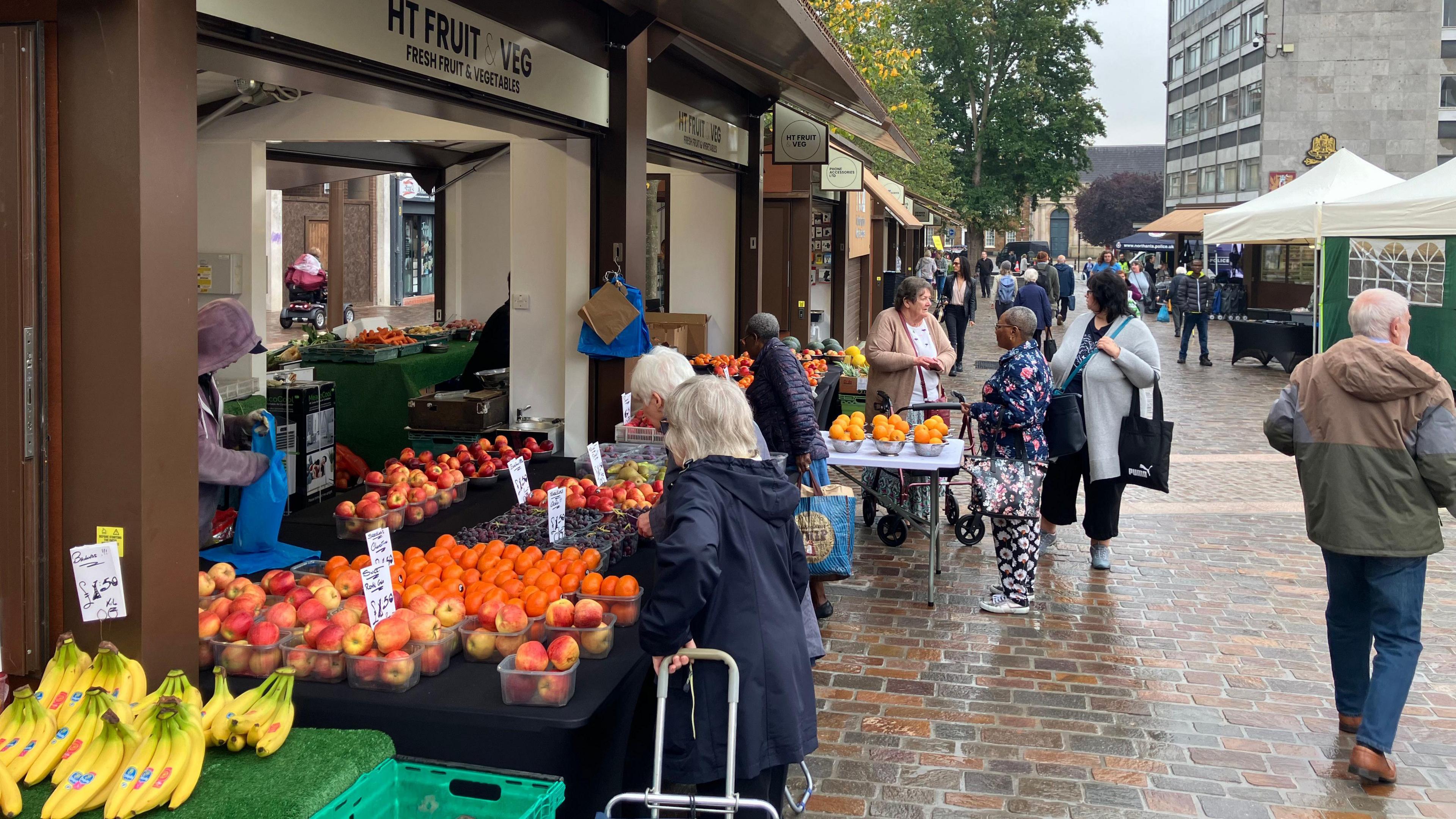 A general shot of people shopping in the new market square area. Two women with white hair look at the wares of a fruit seller, taking a close look at a punnet of oranges. Behind them, two women queue at another fruit seller's stall. 