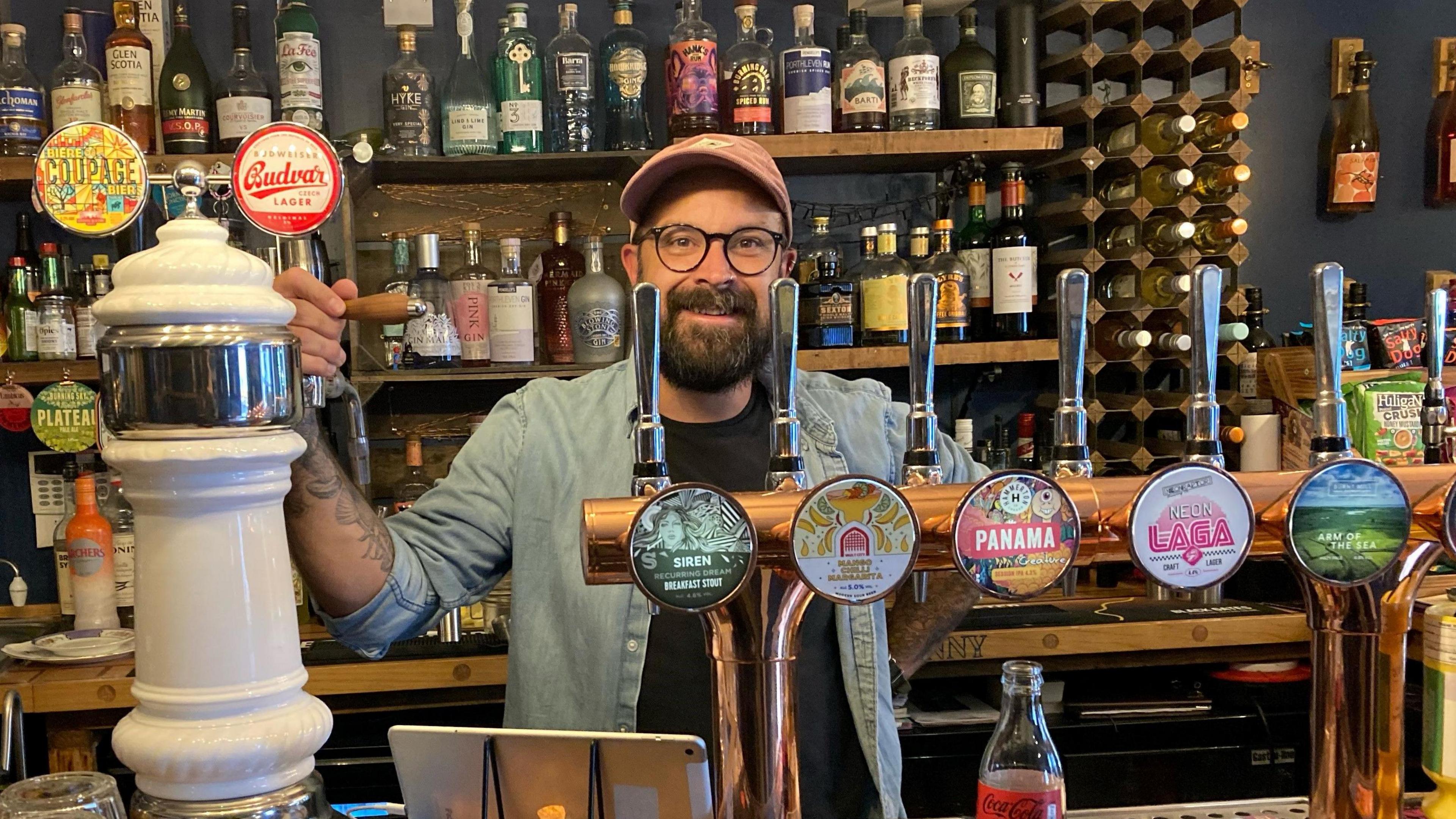 Jamie Stapleton owner of The Tuppenny pub. He is stood behind the bar, with one of his hands on a beer tap. He is wearing a pink cap and has glasses and a beard.