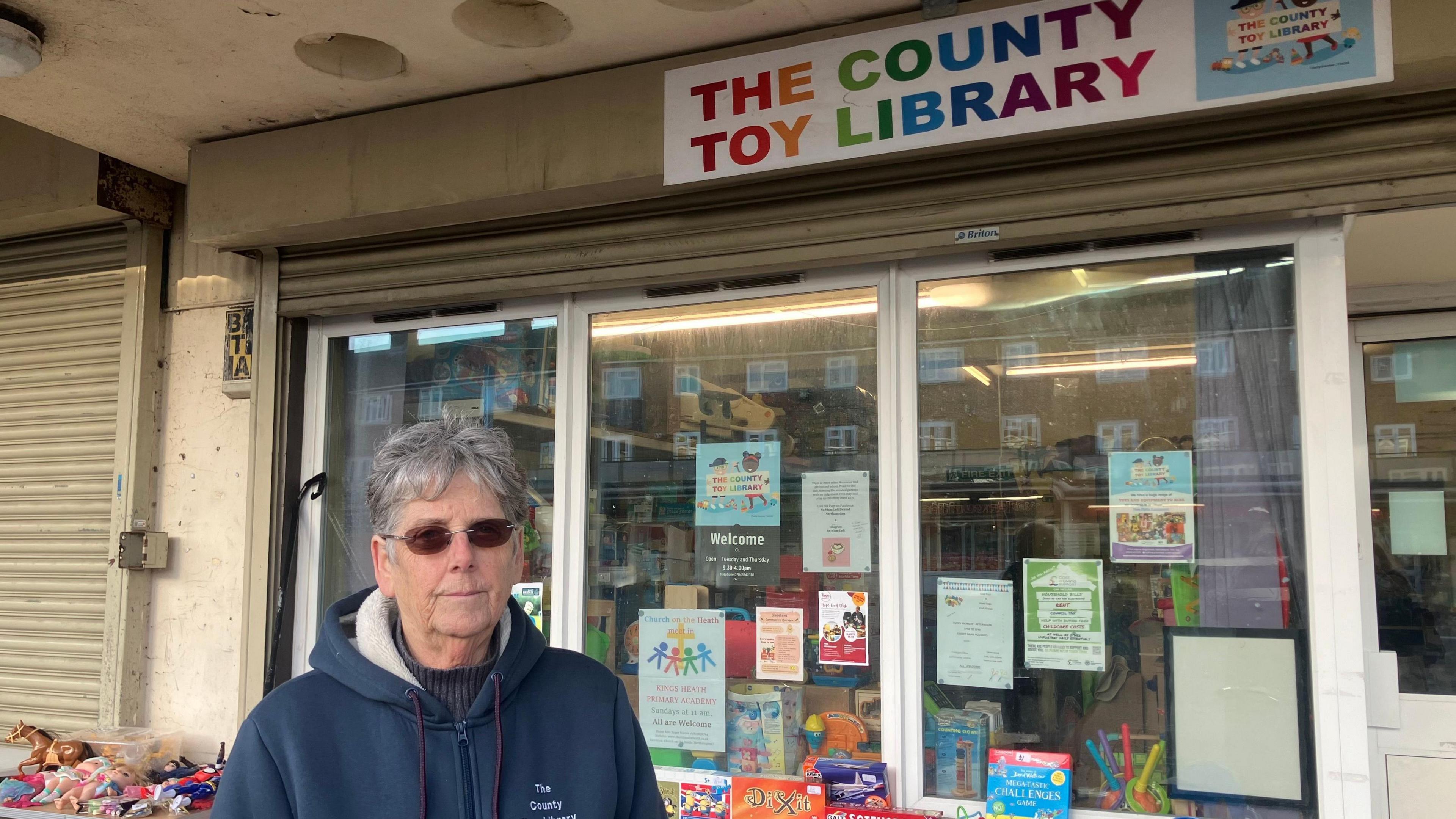 Heather Brakes with short grey hair and dark glasses wearing a blue fleece. She is standing outside the toy library that has various posters in the window and a table outside with books and toys.