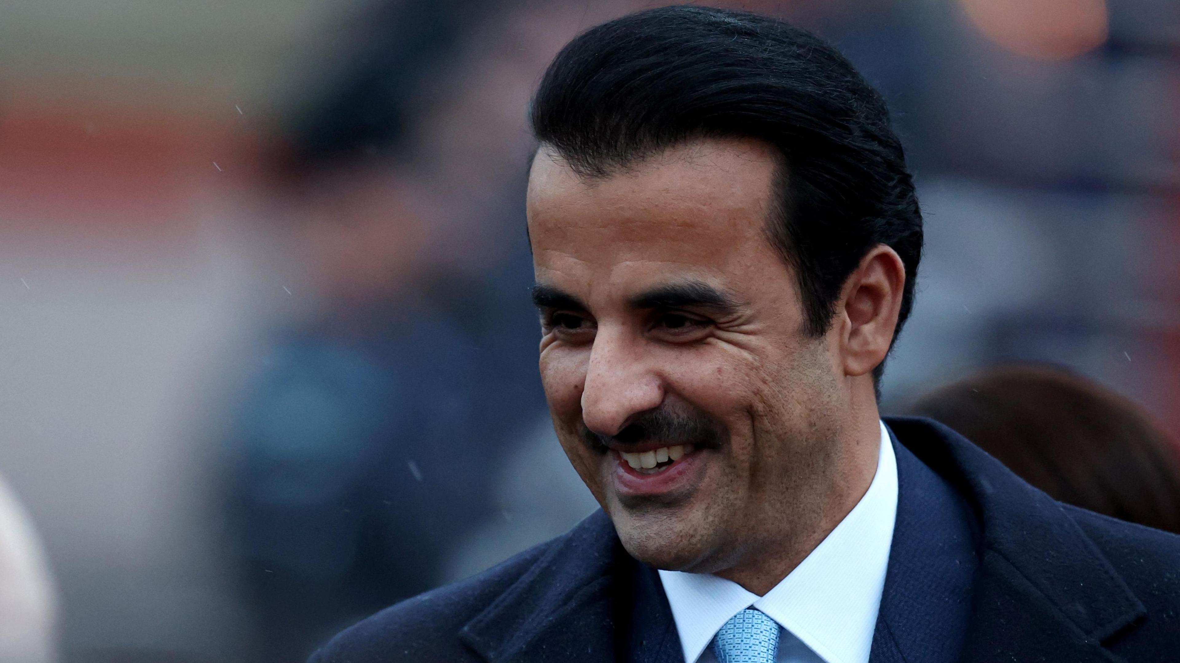 Emir of Qatar smiling while wearing a suit with a light blue tie in the rain. He has short black hair and a black moustache.