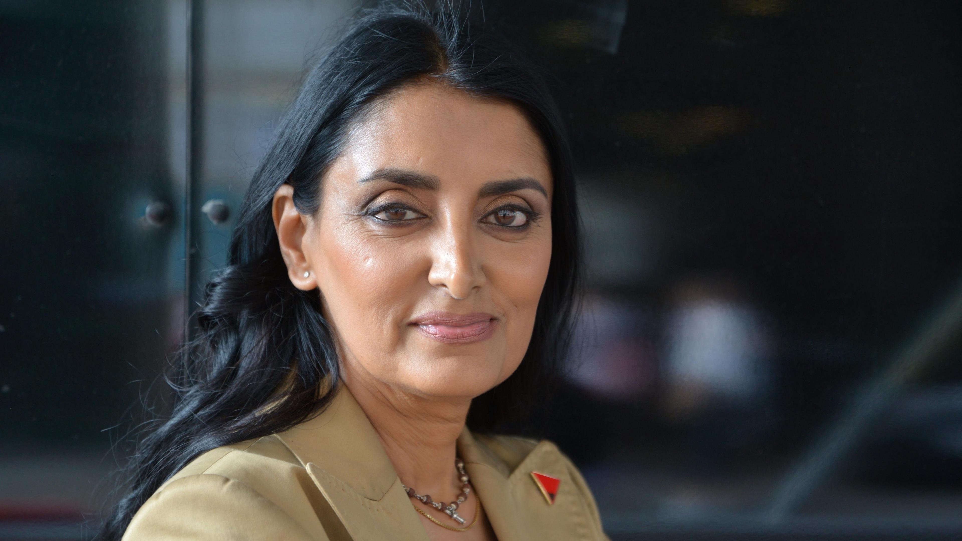 Aneeta Prem looks directly at the camera. She has dark hair and is wearing a pale coloured suit jacket. There is a red and black badge on her lapel in the shape of a triangle. 