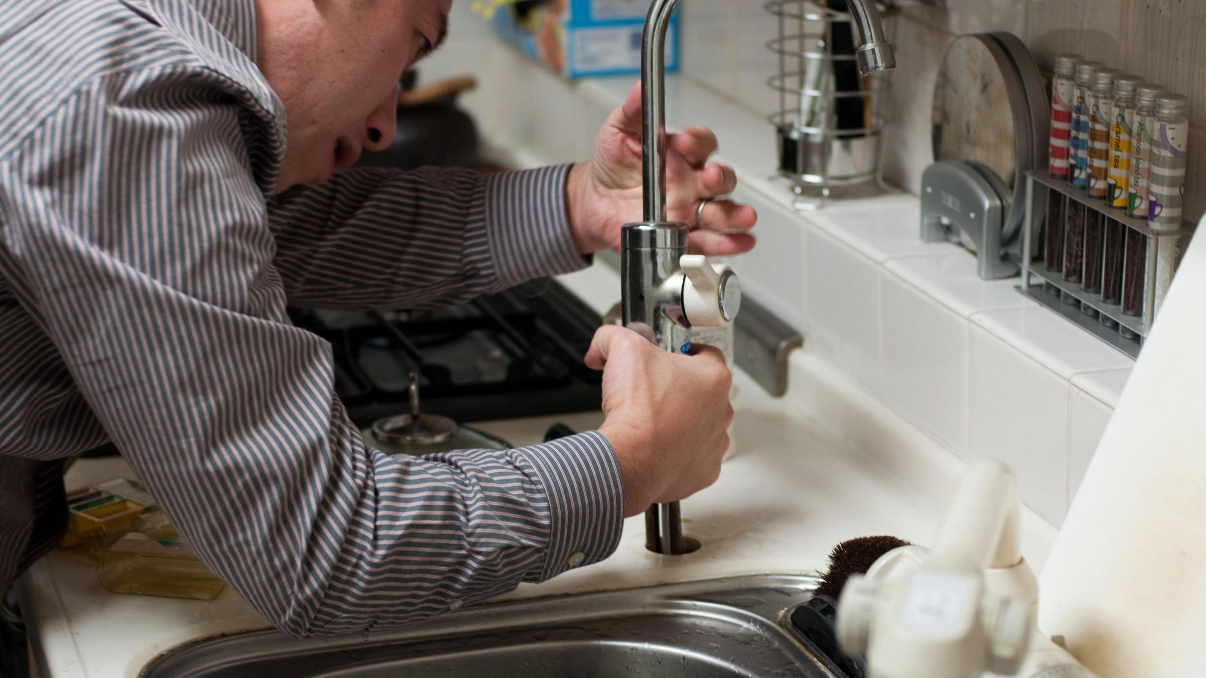 a man wearing a striped shirt is leaning over a chrome kitchen sink. He is holding the base of a chrome kitchen sink tap. He looks as if there is a problem and he is trying to repair it.