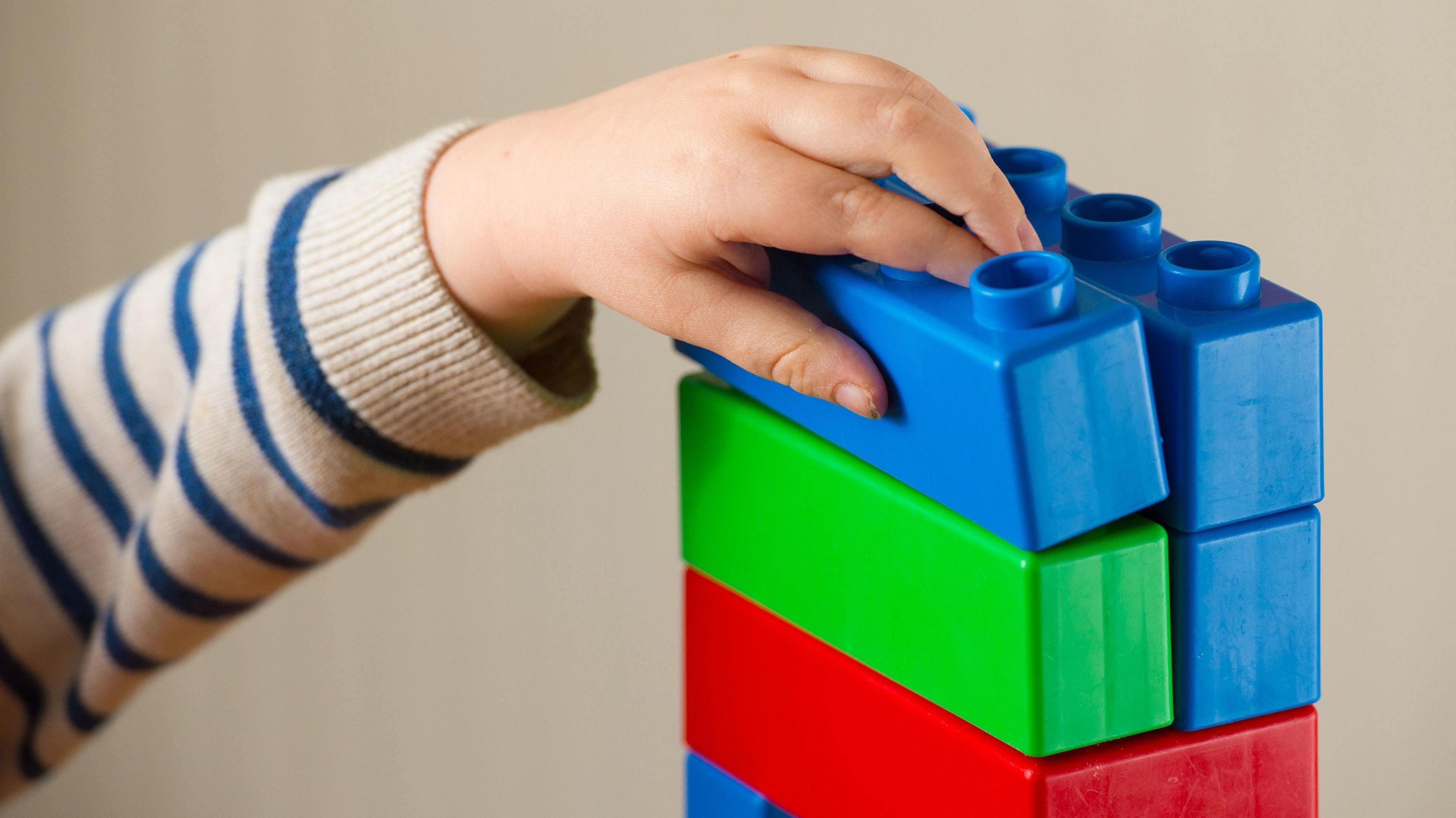 A child's hand reaches for toy building blocks. The child, wearing a grey sweatshirt with blue horizontal stripes, is placing a blue block on top of green red and blue blocks. The wall behind is a neutral colour.