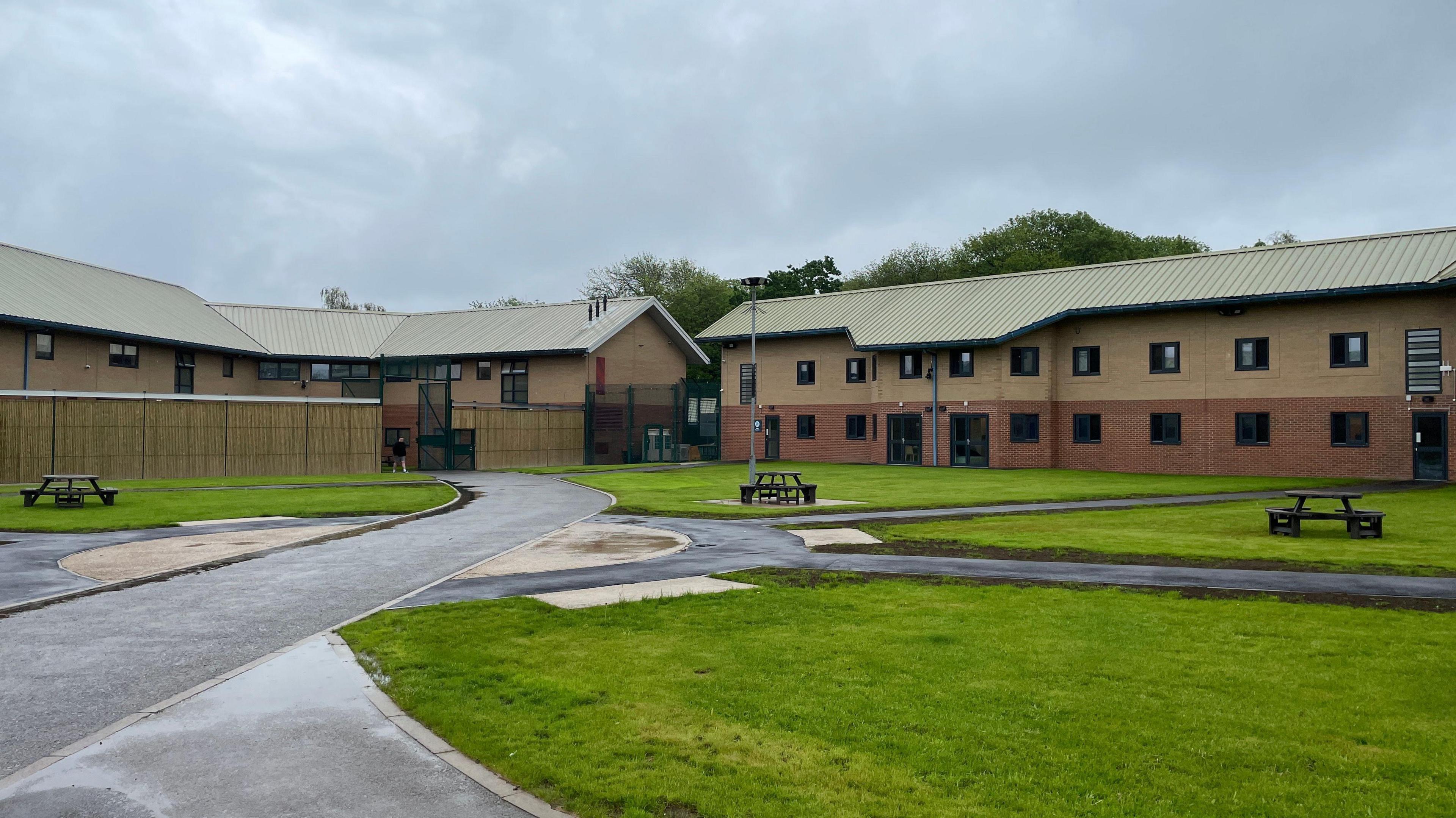 A modern-looking brown building, with a tarmac road leading up to it. In the foreground are areas of grass, with benches 