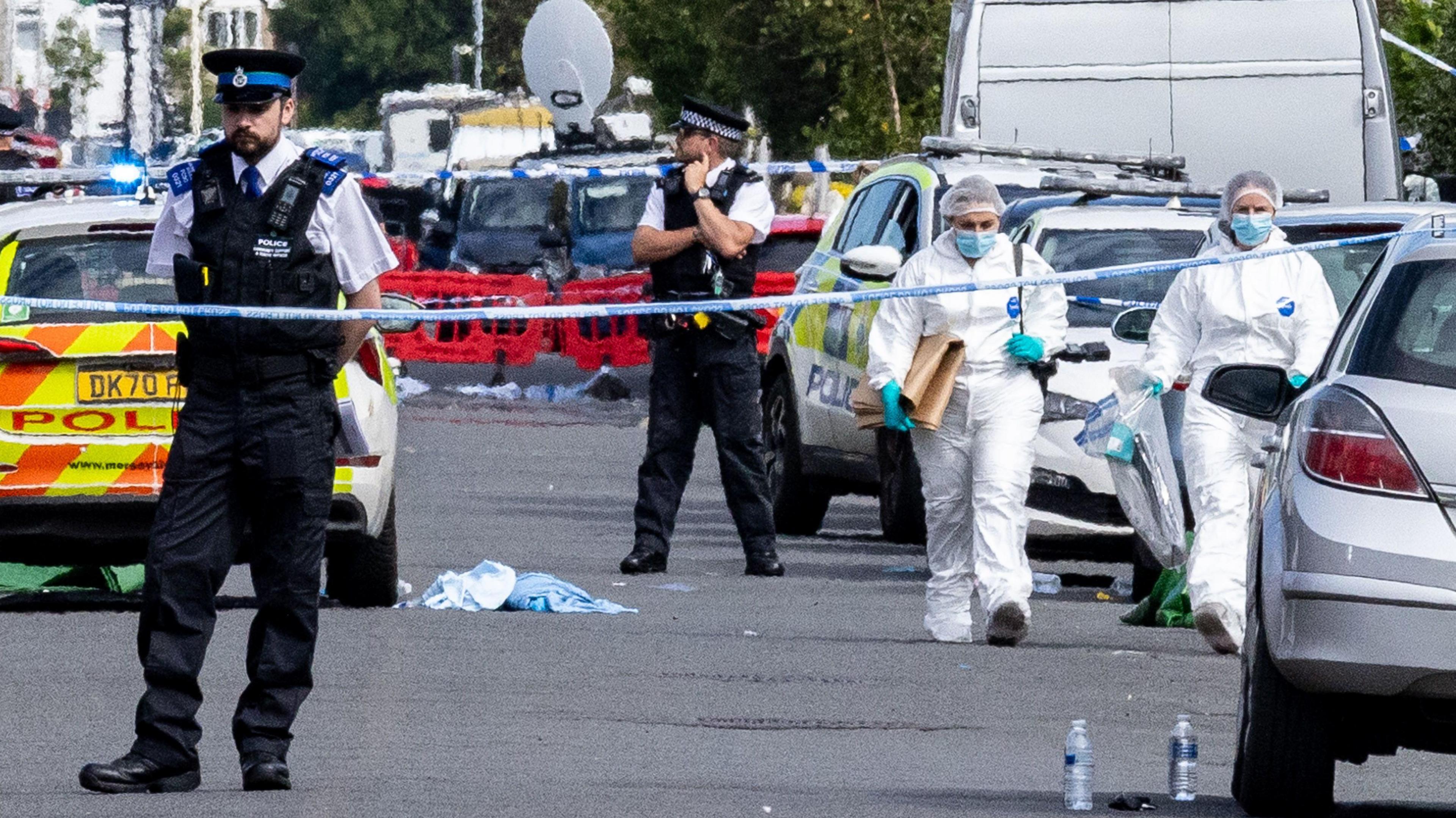 Police officers stand in a road filled with police vans and cars with debris strewn across it behind blue-and-white police tape.