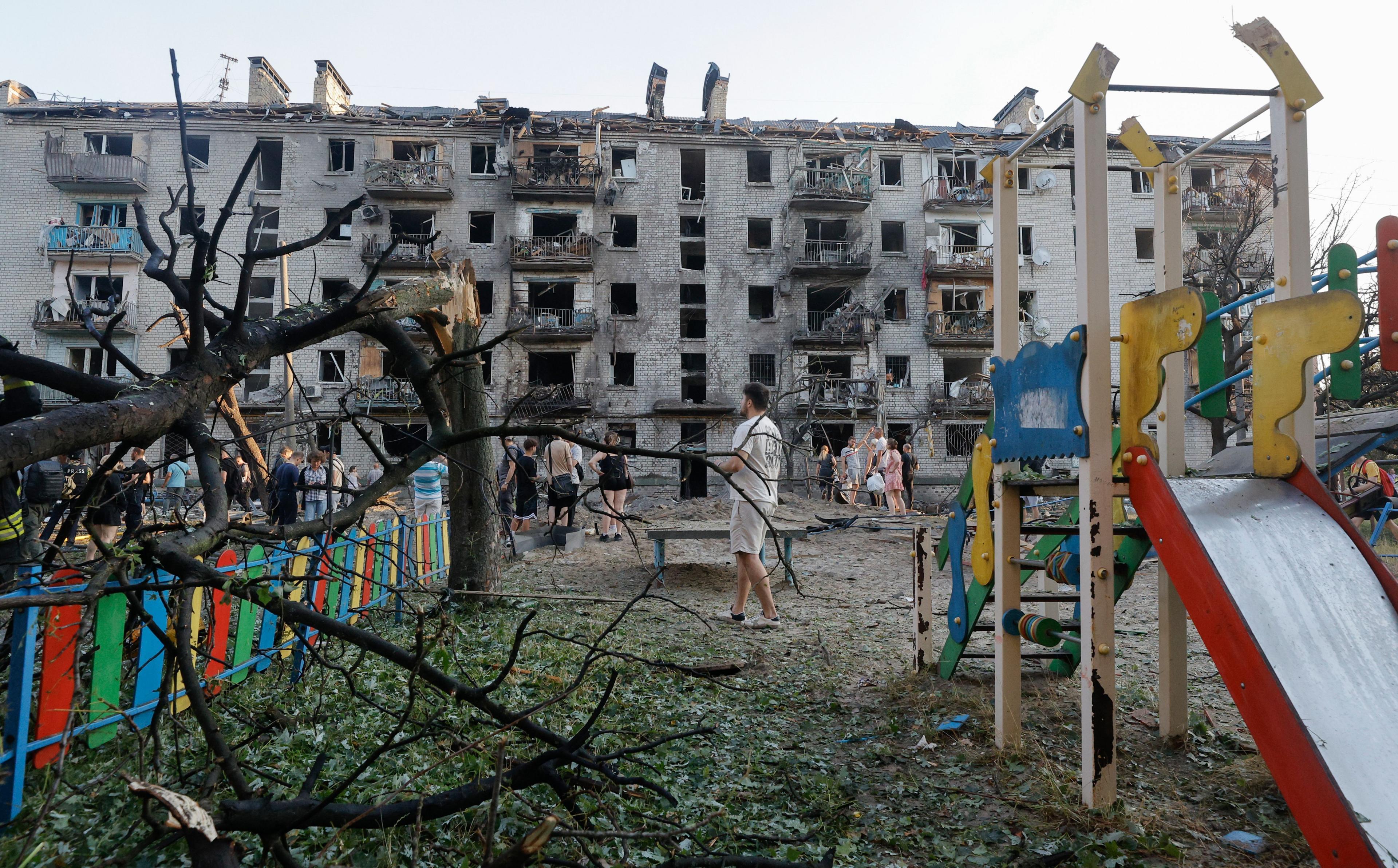 A partially-damaged playground slide in yellow, red and green stands on the right of the frame. A tree split at the trunk is bent over a fence also painted in red, green, yellow and blue. In the background tens of residents walk around the while a partially-destroyed building sits in the background.