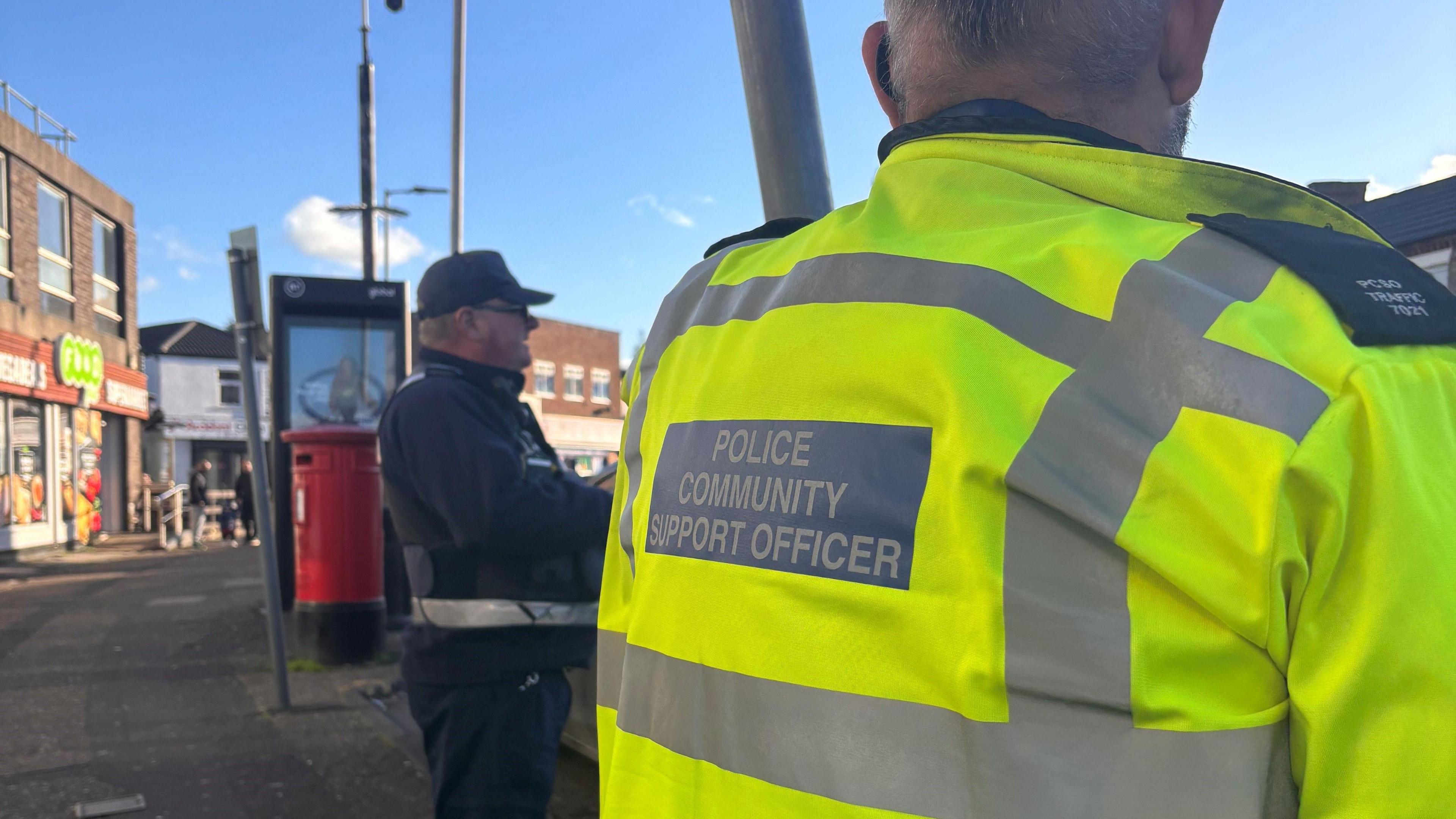 A community support officer with his back to the camera. He wears a high-visibility coat and is stood outside during the day. 